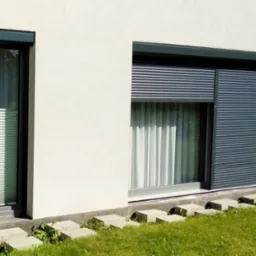 Elewacja budynku z dwoma oknami: w jednym plisy, w drugim roleta zewnętrzna częściowo zasłonięta. Widoczne betonowe obrzeża i trawa.