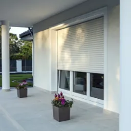 Elegancki, jasny taras z białymi kolumnami i oknem z częściowo opuszczoną roletą zewnętrzną. Kwiaty w donicach dodają uroku.