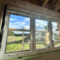 ZASŁOŃ TO EMILIA KOPROWSKA - Trzyczęściowe okno PCV z widokiem na zewnątrz, zamontowane w surowym wnętrzu. Widoczne detale montażowe i krajobraz za oknem z rusztowaniem.