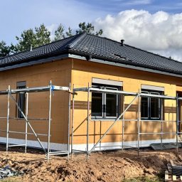ZASŁOŃ TO EMILIA KOPROWSKA - Dom w trakcie budowy z zamontowanymi oknami z roletami zewnętrznymi, widoczne rusztowania i materiały budowlane. Dach pokryty dachówką ceramiczną.
