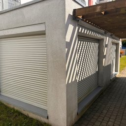 ZASŁOŃ TO EMILIA KOPROWSKA - Zamknięte rolety zewnętrzne w jasnym odcieniu szarości, zamontowane w budynku z szarą elewacją. Cień drewnianej konstrukcji tworzy wzór na roletach po prawej stronie.