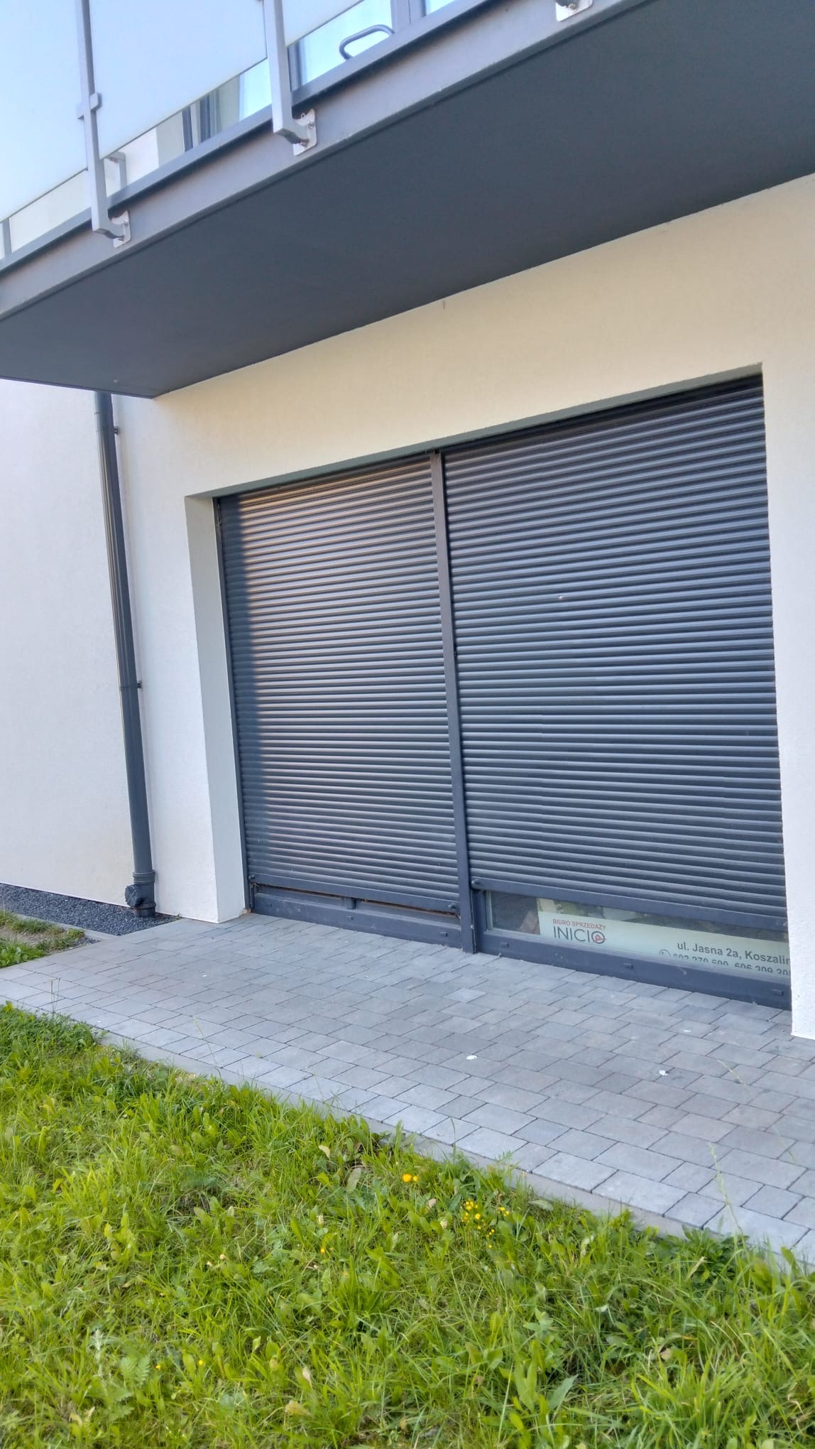 Zewnętrzna roleta antywłamaniowa w kolorze grafitowym, zamontowana w nowoczesnym budynku z jasną elewacją. Widoczny fragment chodnika z kostki brukowej i trawnika.