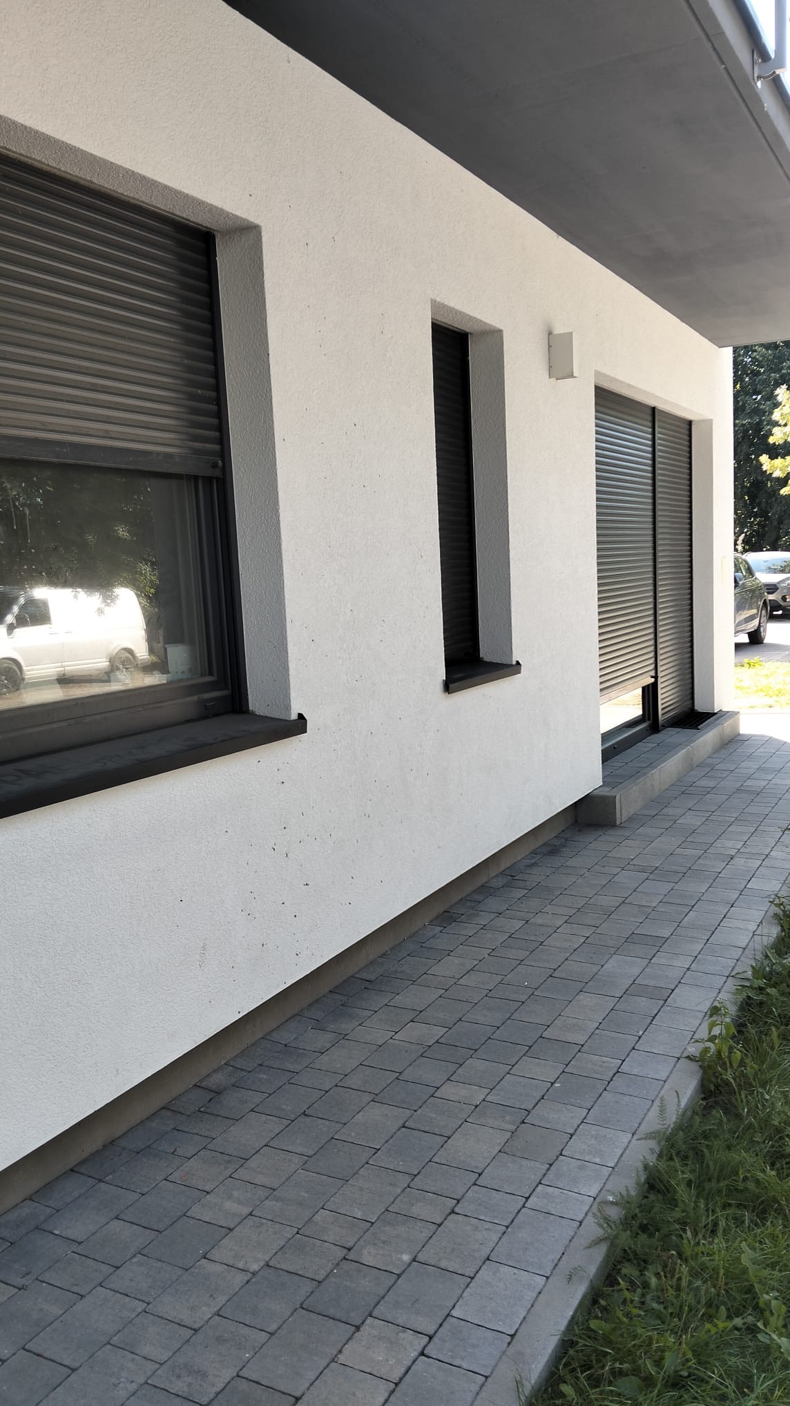 Elewacja domu z dwoma oknami z roletami zewnętrznymi w kolorze antracytowym. Widoczne szare płytki chodnikowe i fragment trawnika.