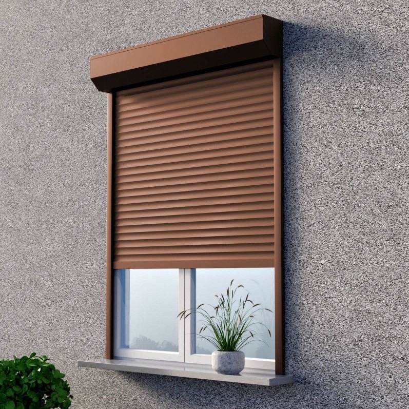 Brązowa roleta zewnętrzna częściowo opuszczona na oknie budynku z szarą elewacją. Na parapecie doniczka z trawą.