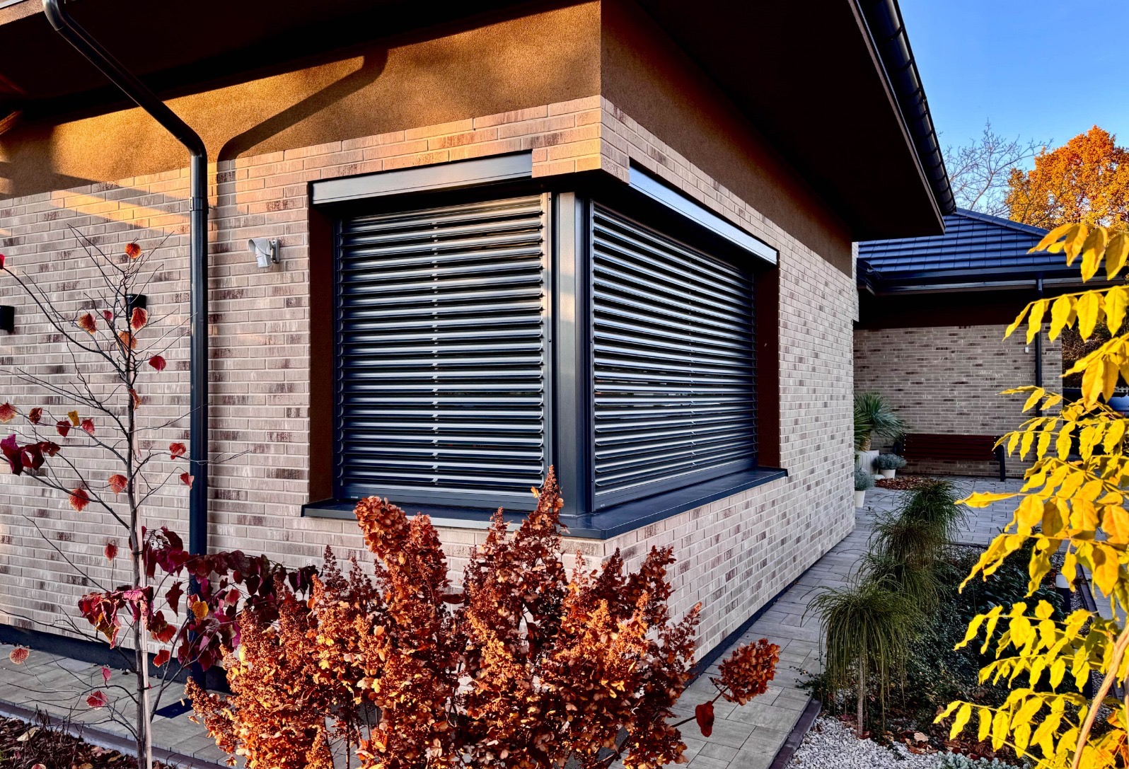 Elewacja domu z cegły klinkierowej z oknem narożnym i zewnętrznymi żaluzjami aluminiowymi w kolorze grafitowym, widok z poziomu ogrodu jesienią.