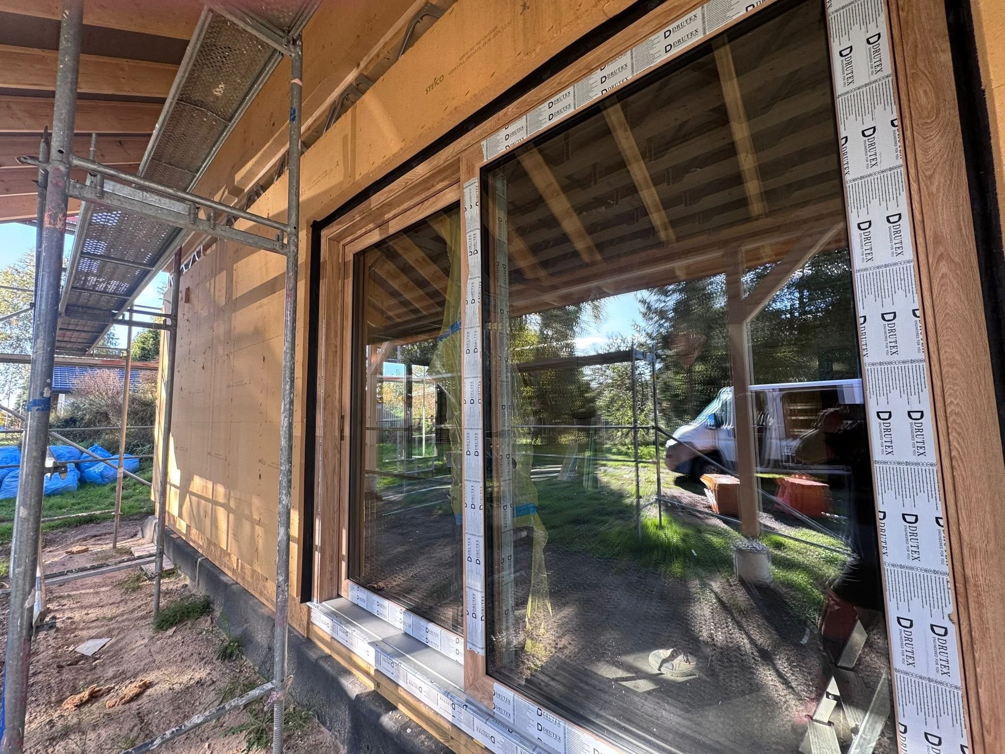 Montaż okna z drewnianą ramą w surowym budynku. Widoczne rusztowanie i folia ochronna na profilach. W szybie odbija się otoczenie.