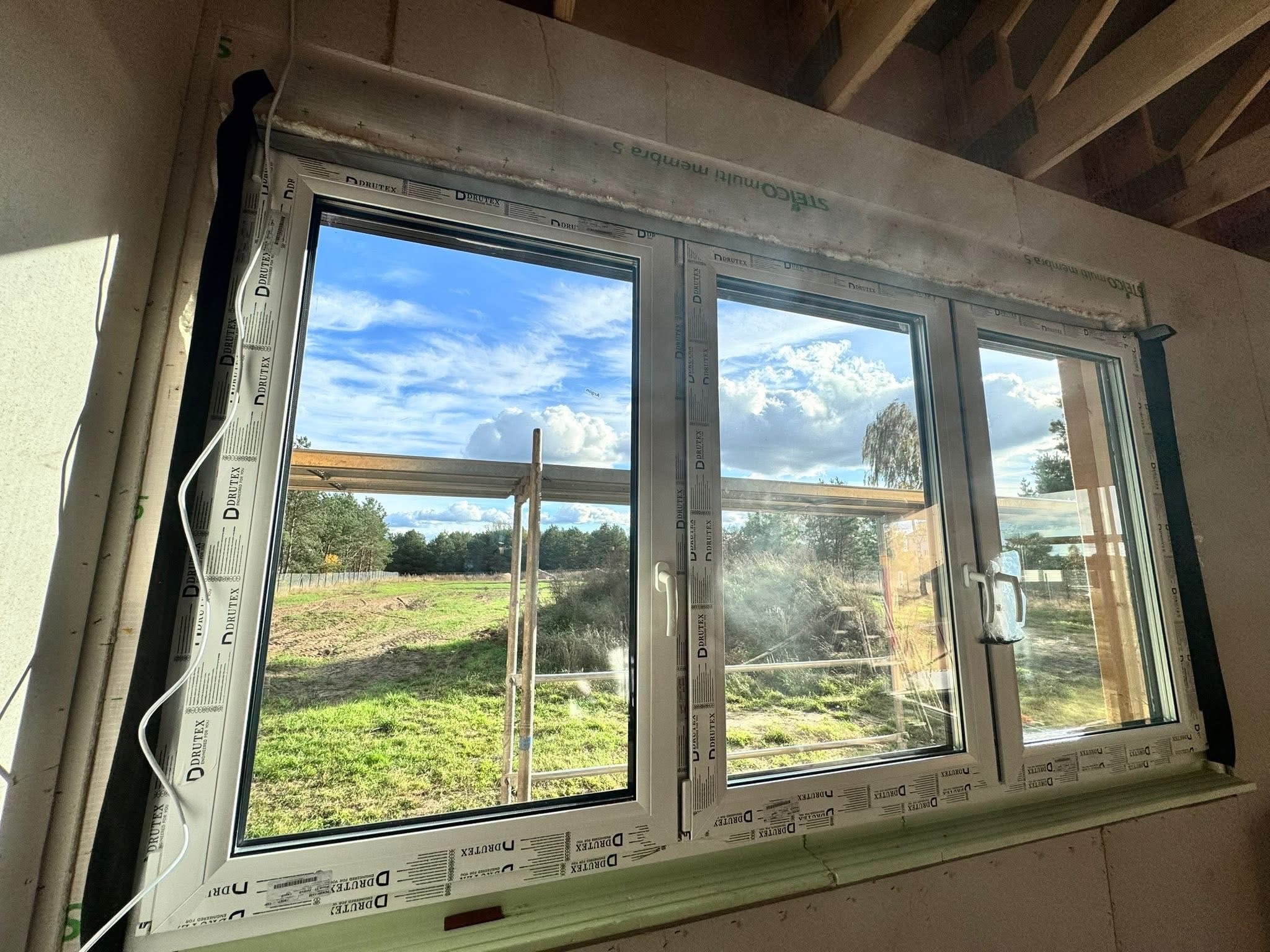 Trzyczęściowe okno PCV z widokiem na zewnątrz, zamontowane w surowym wnętrzu. Widoczne detale montażowe i krajobraz za oknem z rusztowaniem.