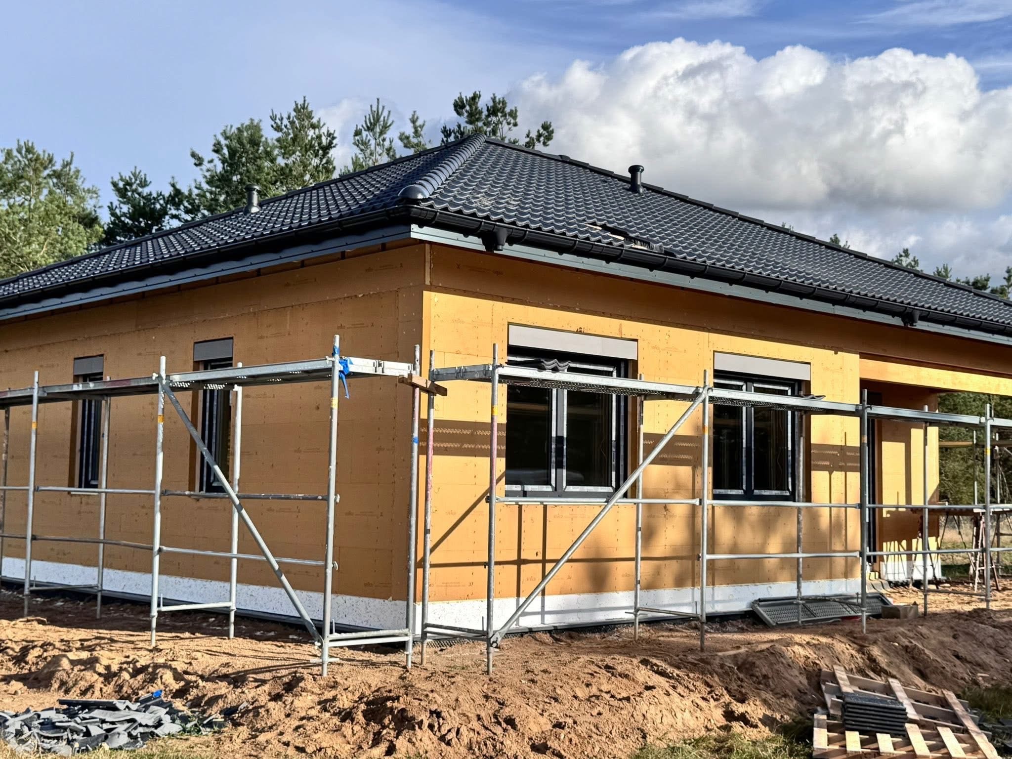 Dom w trakcie budowy z zamontowanymi oknami z roletami zewnętrznymi, widoczne rusztowania i materiały budowlane. Dach pokryty dachówką ceramiczną.