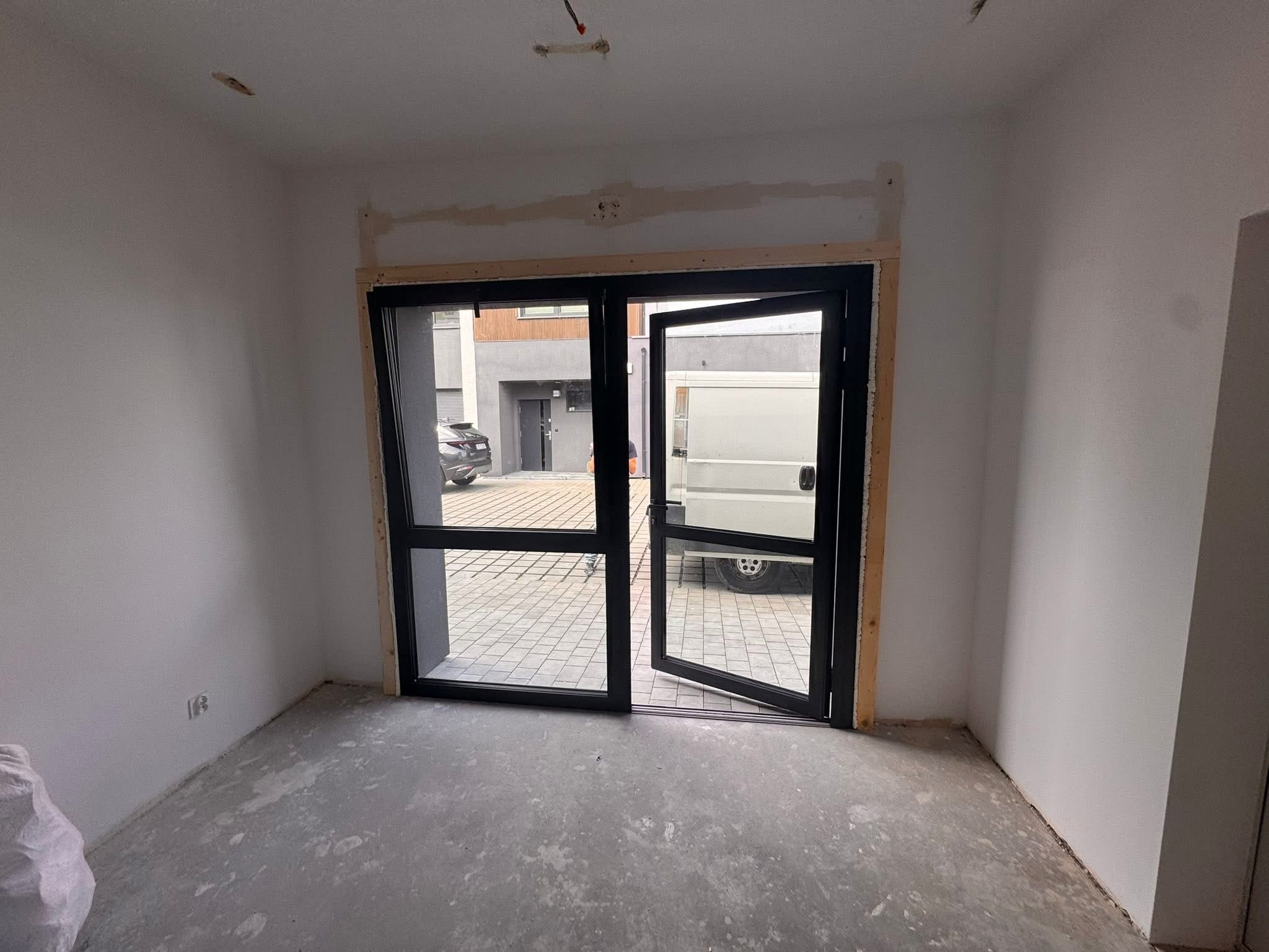Nowoczesne, czarne drzwi balkonowe z widokiem na podjazd i budynek. Jedno skrzydło otwarte, surowy stan wykończenia wnętrza, widoczne ślady po montażu.