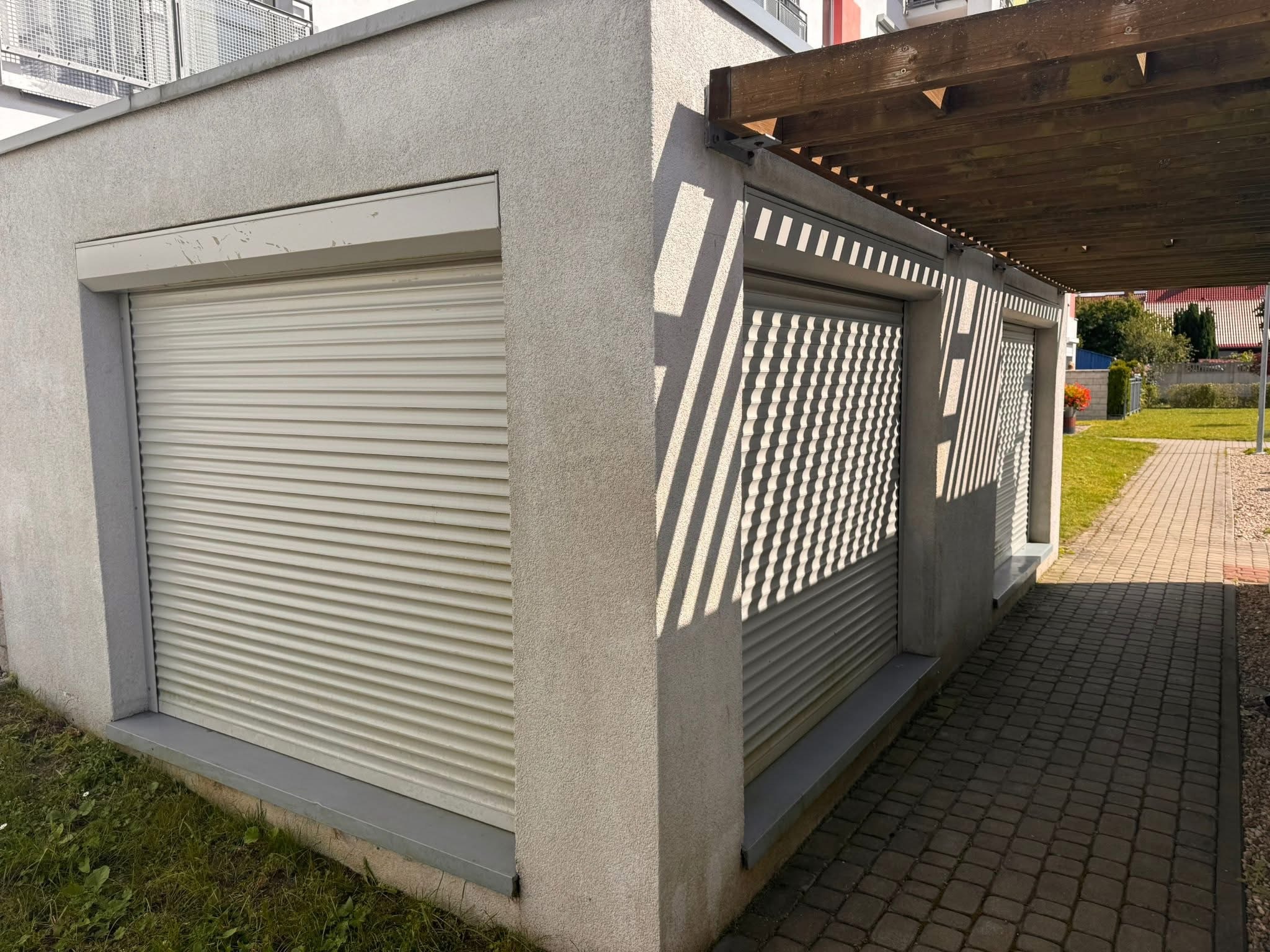 Zamknięte rolety zewnętrzne w jasnym odcieniu szarości, zamontowane w budynku z szarą elewacją. Cień drewnianej konstrukcji tworzy wzór na roletach po prawej stronie.