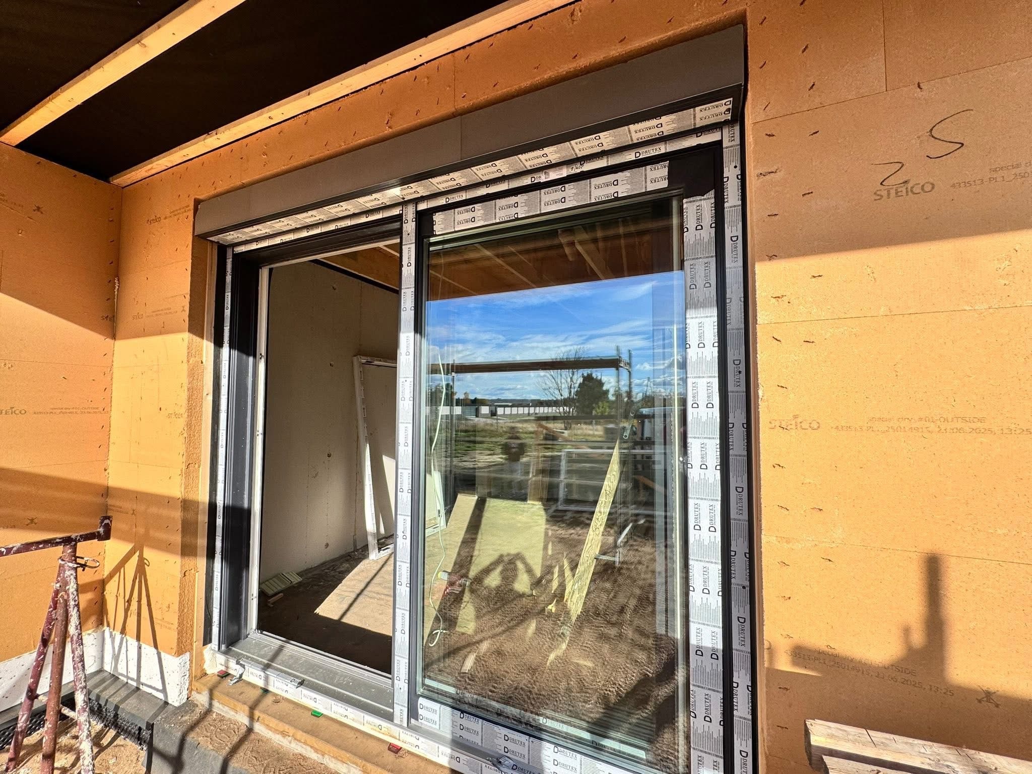 Montaż okna przesuwnego z roletą zewnętrzną w surowym stanie. Widoczne materiały izolacyjne i rusztowanie. Okno z szybami odblaskowymi.