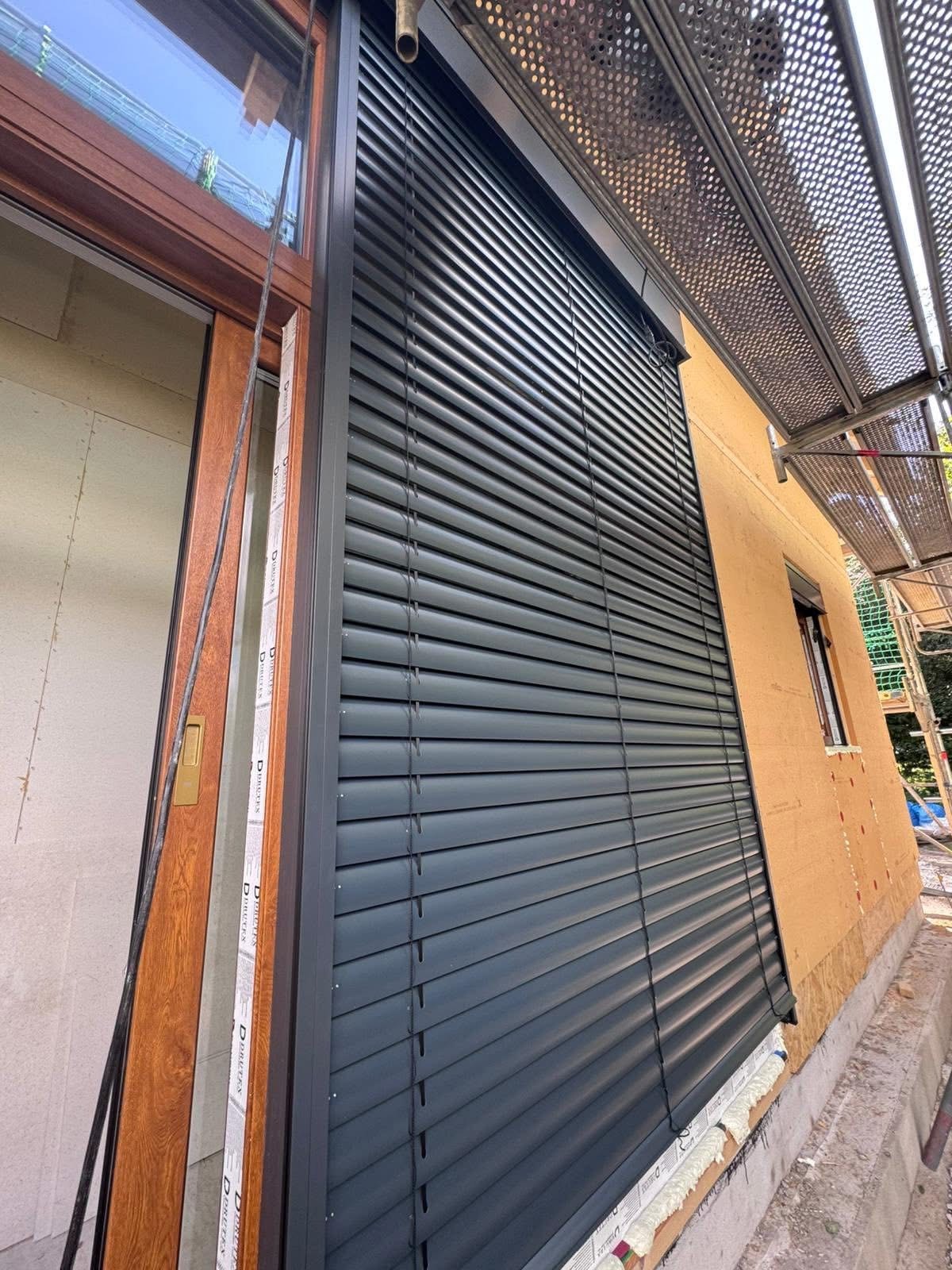 Eleganckie, grafitowe rolety zewnętrzne zamontowane na elewacji budynku w trakcie wykończenia. Widoczne rusztowania i okno z drewnianą ramą.