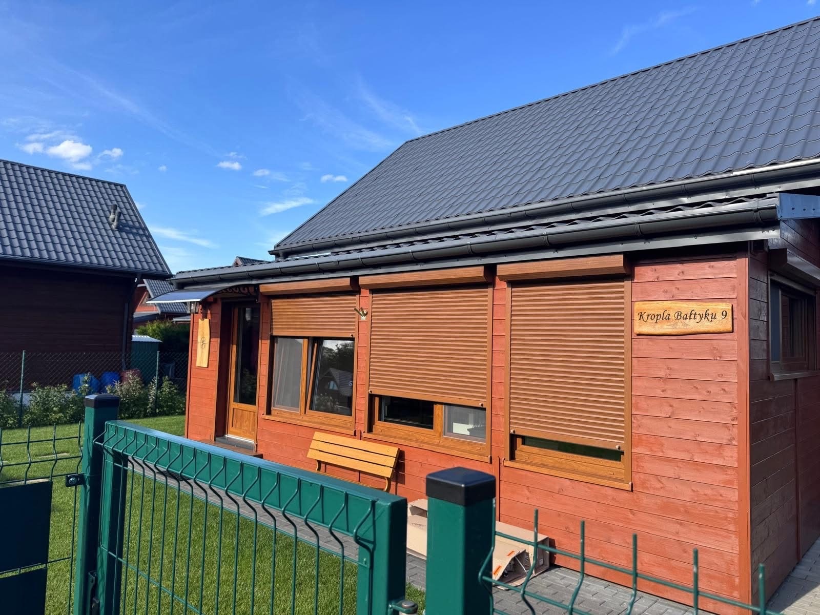 Drewniany domek letniskowy z brązowymi roletami zewnętrznymi, częściowo opuszczonymi. Widoczny fragment zielonego ogrodzenia i tabliczka z adresem.