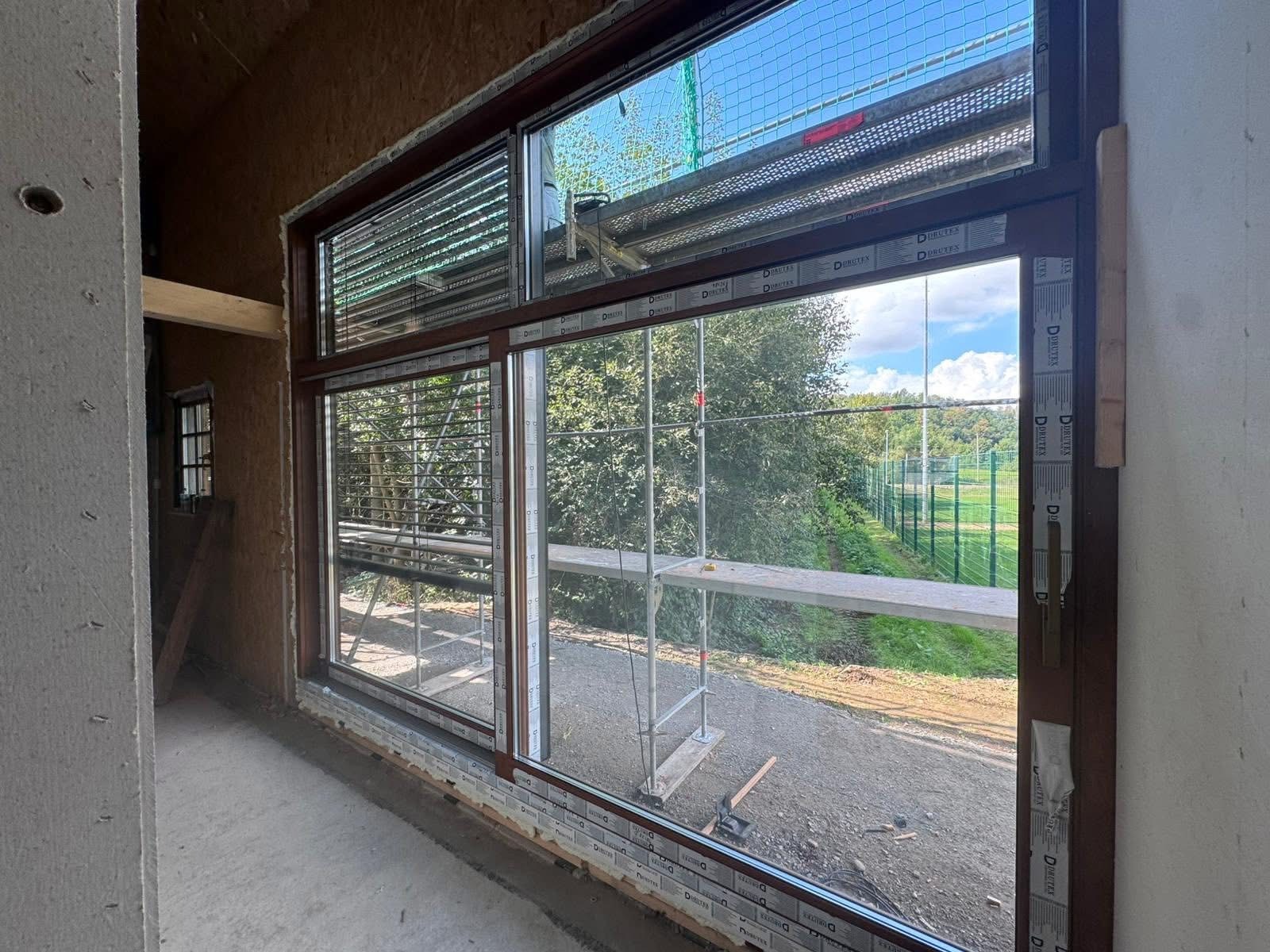 Montaż okna z brązową ramą w surowym wnętrzu. Widok na zewnątrz z rusztowaniem i zielenią. Okno przesuwne z żaluzjami. Detal montażu.