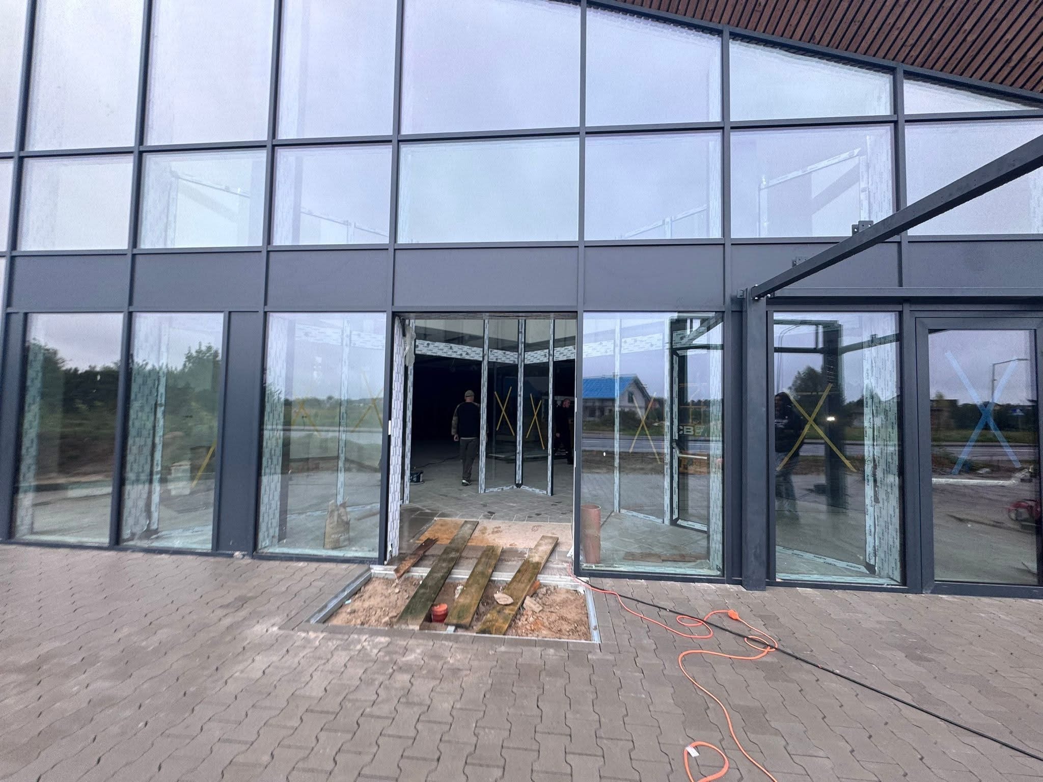 Montaż okien aluminiowych w budynku komercyjnym. Widoczne ramy okienne, częściowo zamontowane szyby i przygotowane podłoże. Prace wykończeniowe w toku.