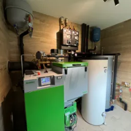 Nowoczesna kotłownia z piecem Bio Compact, zbiornikiem buforowym i osprzętem. Widoczne liczne zawory, manometry i elementy instalacji grzewczej. Ściany wyłożone płytkami imitującymi drewno.