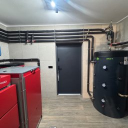 OSAINSTAL Bartosz Osowicki - Kotłownia z piecem Defro, zbiornikiem Drewmet i czarnymi rurami grzewczymi na tle beżowych płytek. Widok na zainstalowany system grzewczy w pomieszczeniu.