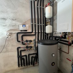 OSAINSTAL Bartosz Osowicki - Złożona instalacja grzewcza z czarnymi rurami, białymi zbiornikami wyrównawczymi i szarym zasobnikiem ciepłej wody na tle surowej, betonowej ściany.