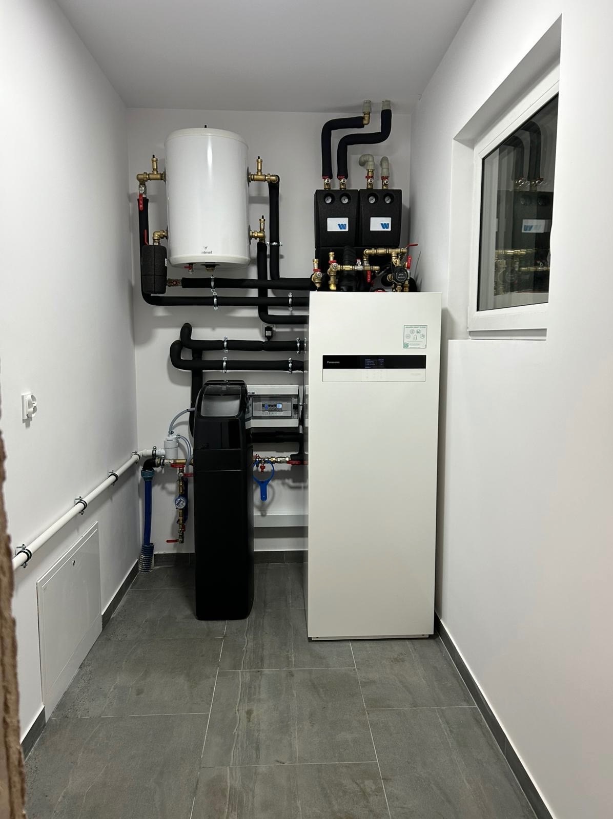 Nowoczesna kotłownia z białym piecem, zasobnikiem wody i czarnymi rurami. Szare płytki podłogowe. Minimalistyczny design w jasnym pomieszczeniu.