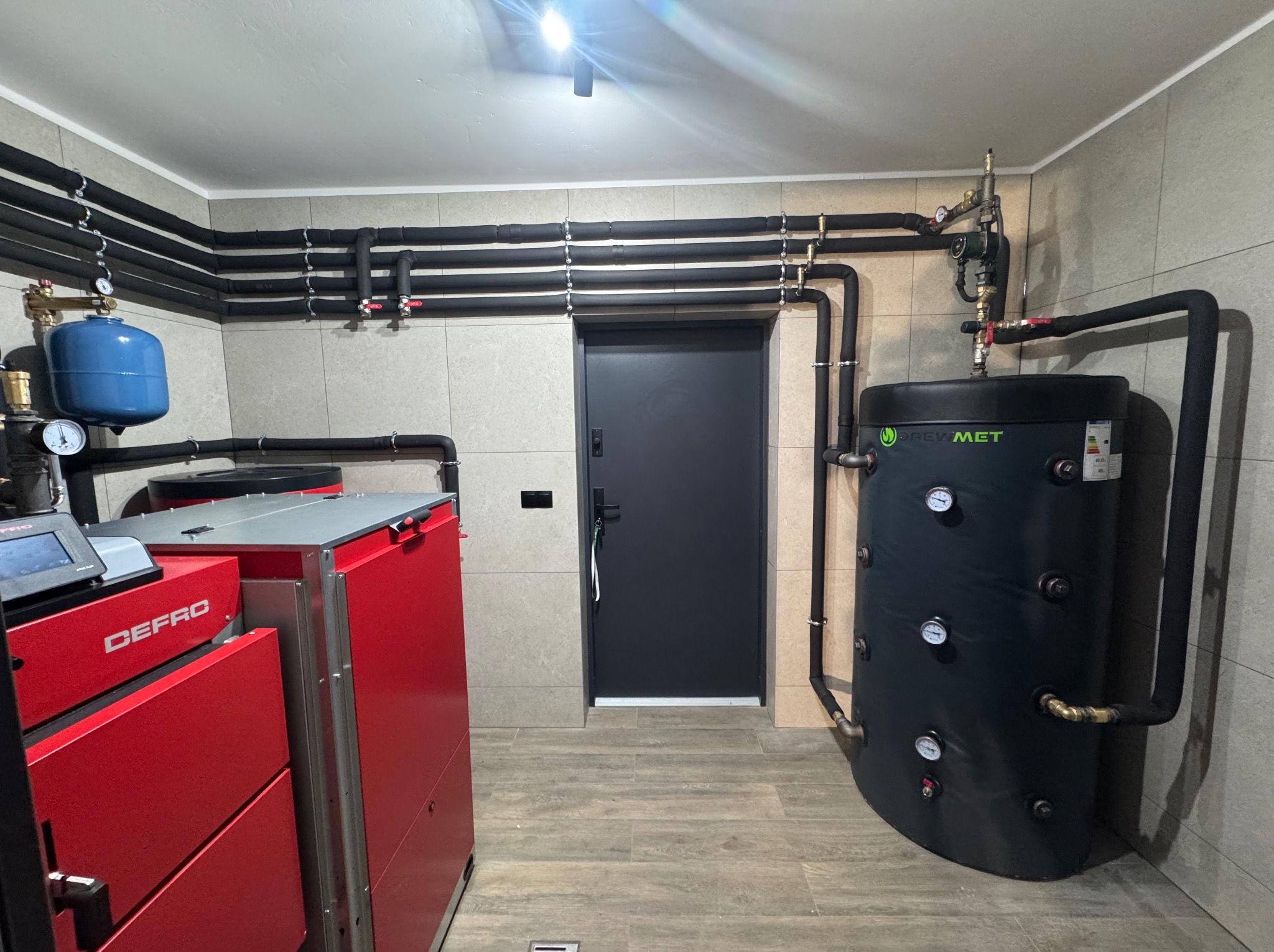 Kotłownia z piecem Defro, zbiornikiem Drewmet i czarnymi rurami grzewczymi na tle beżowych płytek. Widok na zainstalowany system grzewczy w pomieszczeniu.