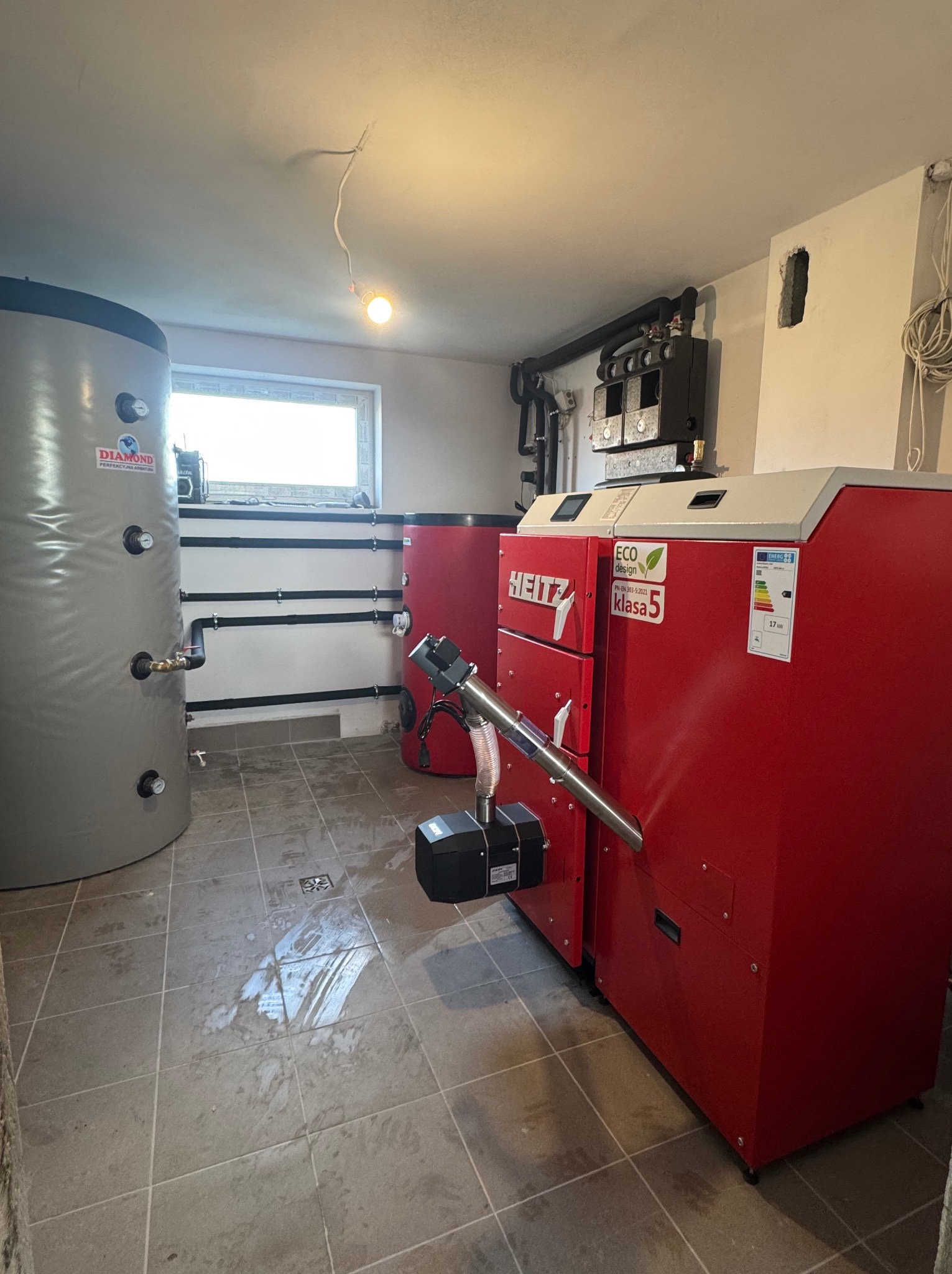 Kotłownia z czerwonym piecem HEITZ Eco Design Klasa 5, szarym zbiornikiem Diamond i czarnymi rurami. Widoczne okno i fragment instalacji elektrycznej.