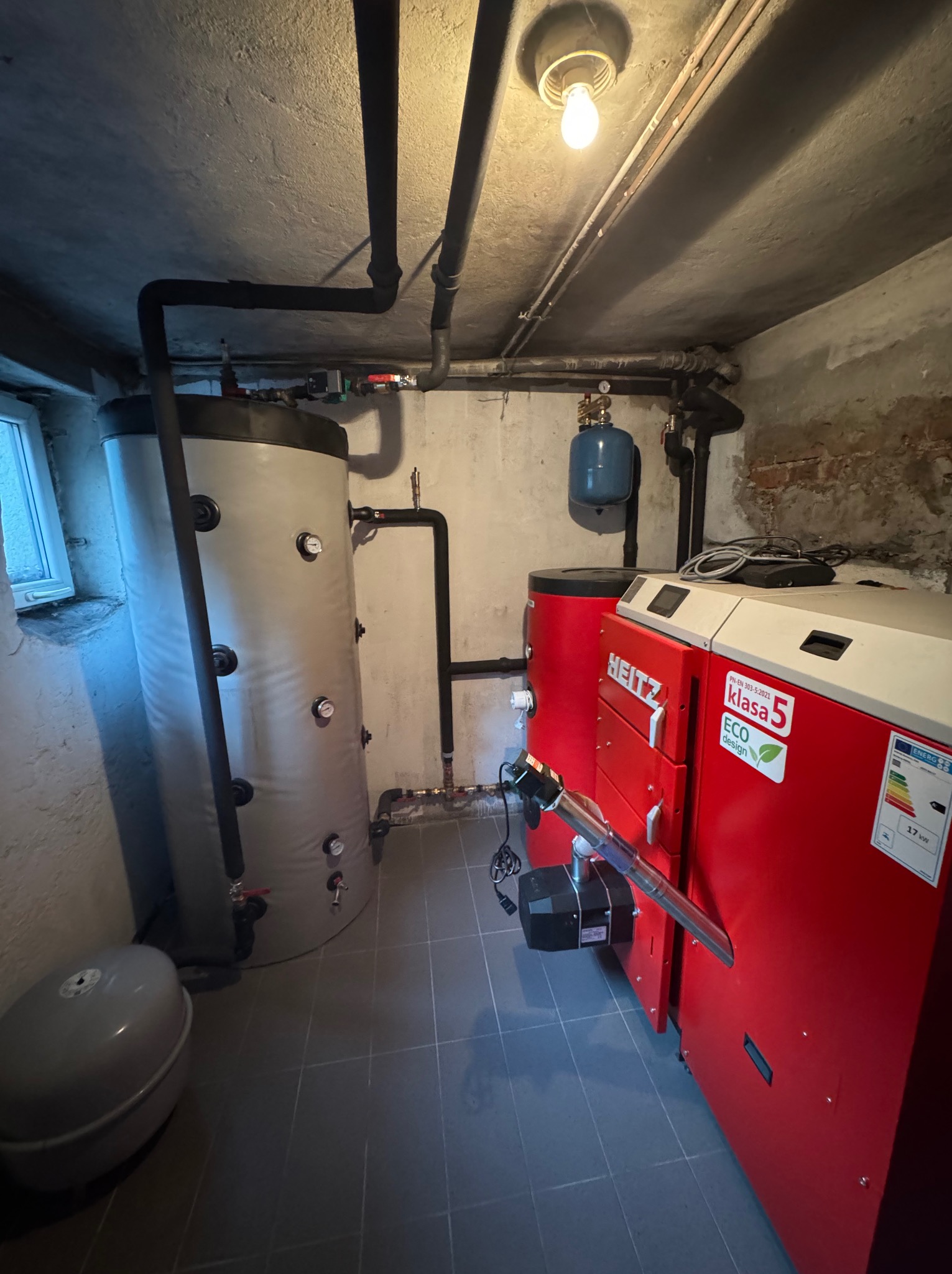 Czerwony piec CO marki Heiz z podajnikiem, obok szary zbiornik akumulacyjny w kotłowni z szarą podłogą i instalacją rurową pod sufitem. Widoczne okno i oświetlenie.