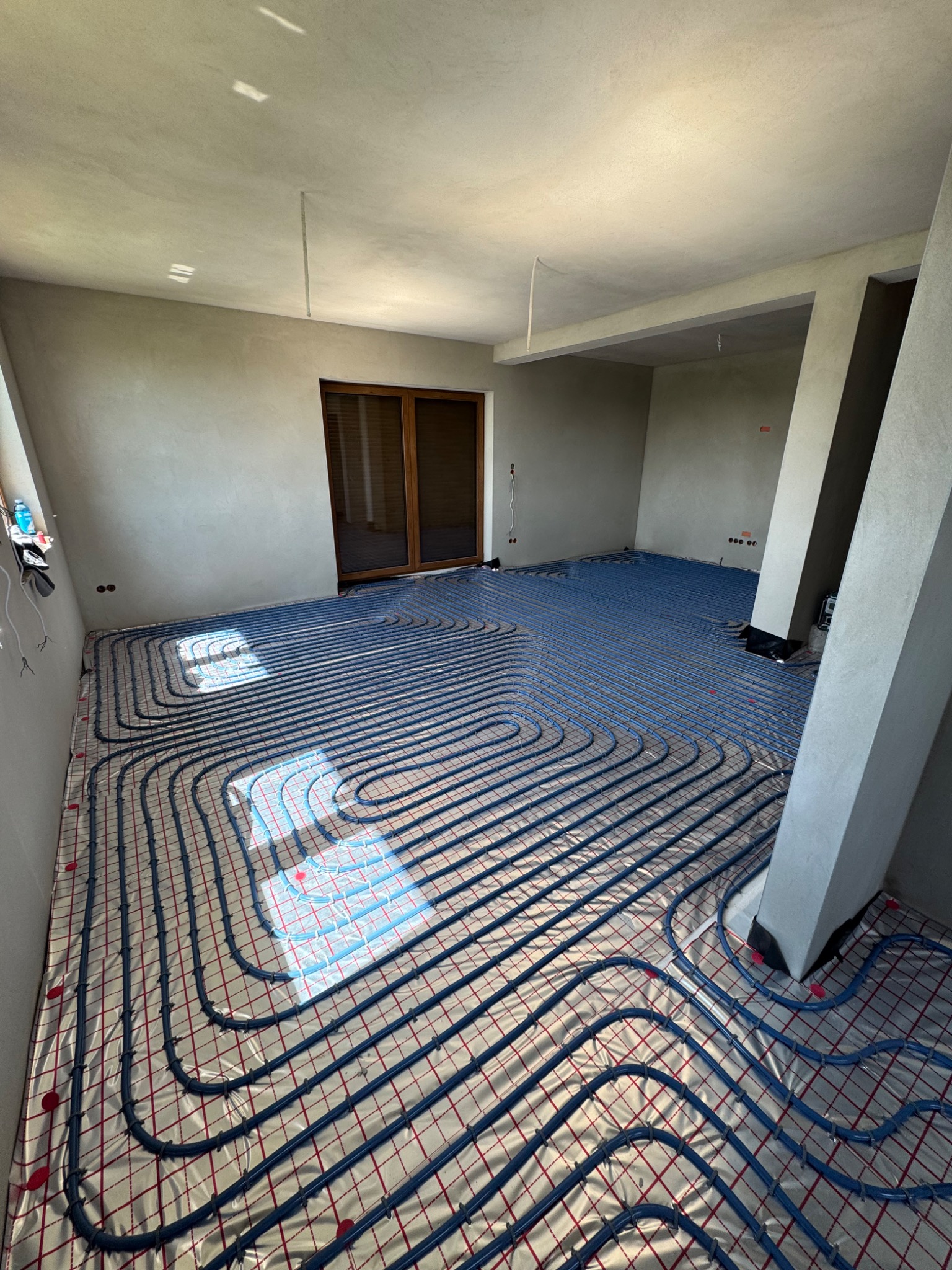 Niebieskie rury ogrzewania podłogowego ułożone na srebrnej folii w surowym wnętrzu z oknem i drzwiami balkonowymi. Widoczne elementy konstrukcyjne.
