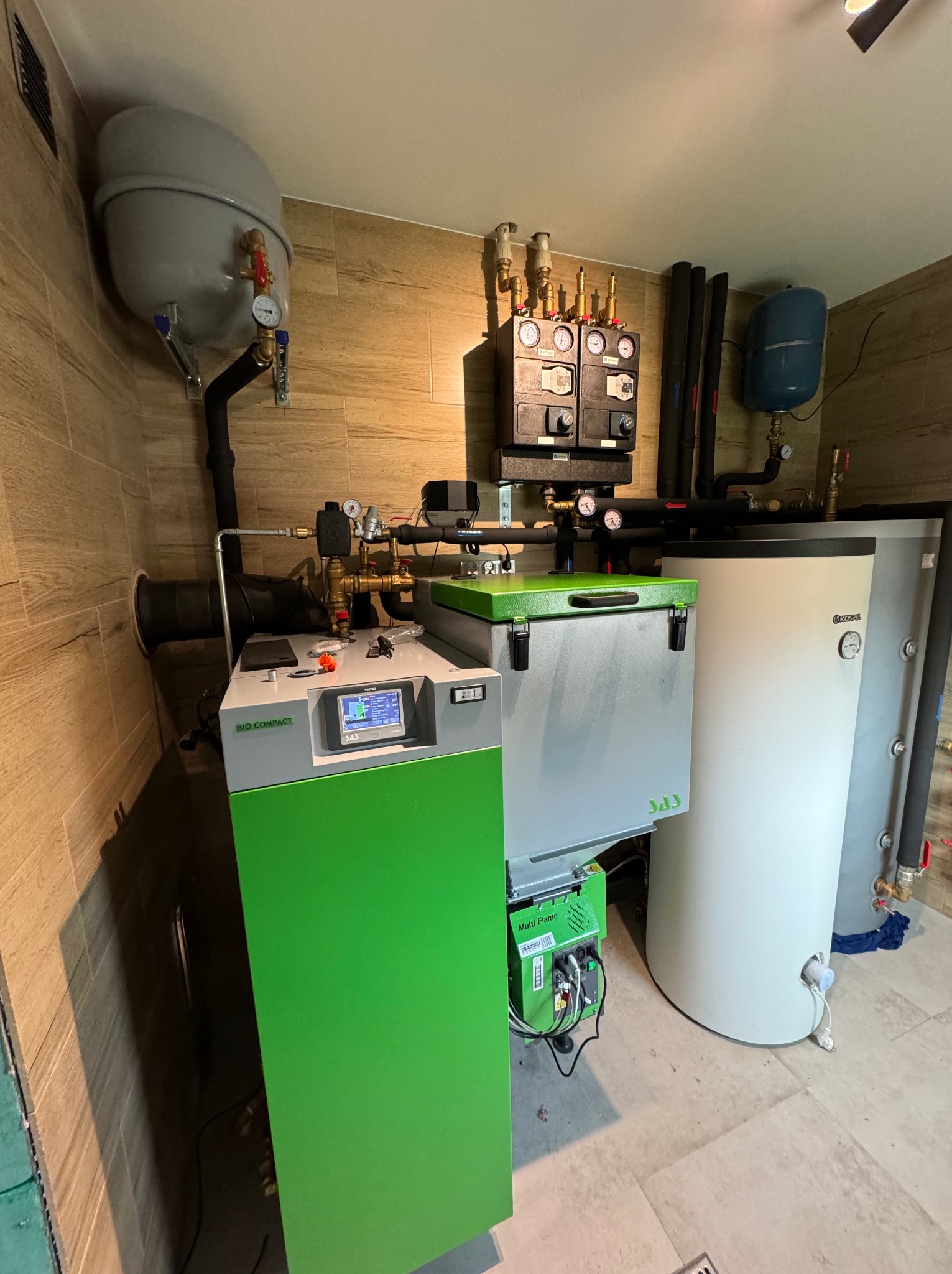 Zestaw urządzeń kotłowni: piec Bio Compact z zielonym panelem, zbiornik Kospel i osprzęt. Widoczne rury, zawory i manometry. Całość w pomieszczeniu wyłożonym płytkami imitującymi drewno.