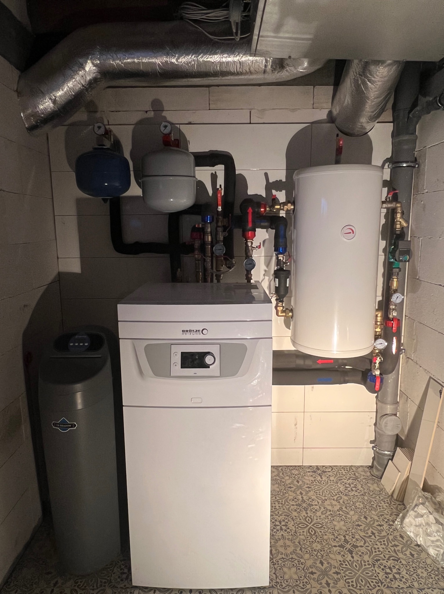 Biały piec CO marki Brötje z szarym zmiękczaczem wody, rurami i zbiornikami wyrównawczymi na tle białych płytek i wzorzystej podłogi w kotłowni.