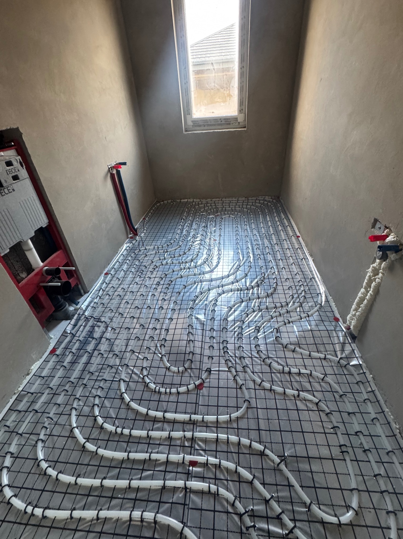 Ułożone rury ogrzewania podłogowego na czarnej siatce w surowym wnętrzu z oknem dachowym. Widoczne elementy instalacji sanitarnej na ścianach.