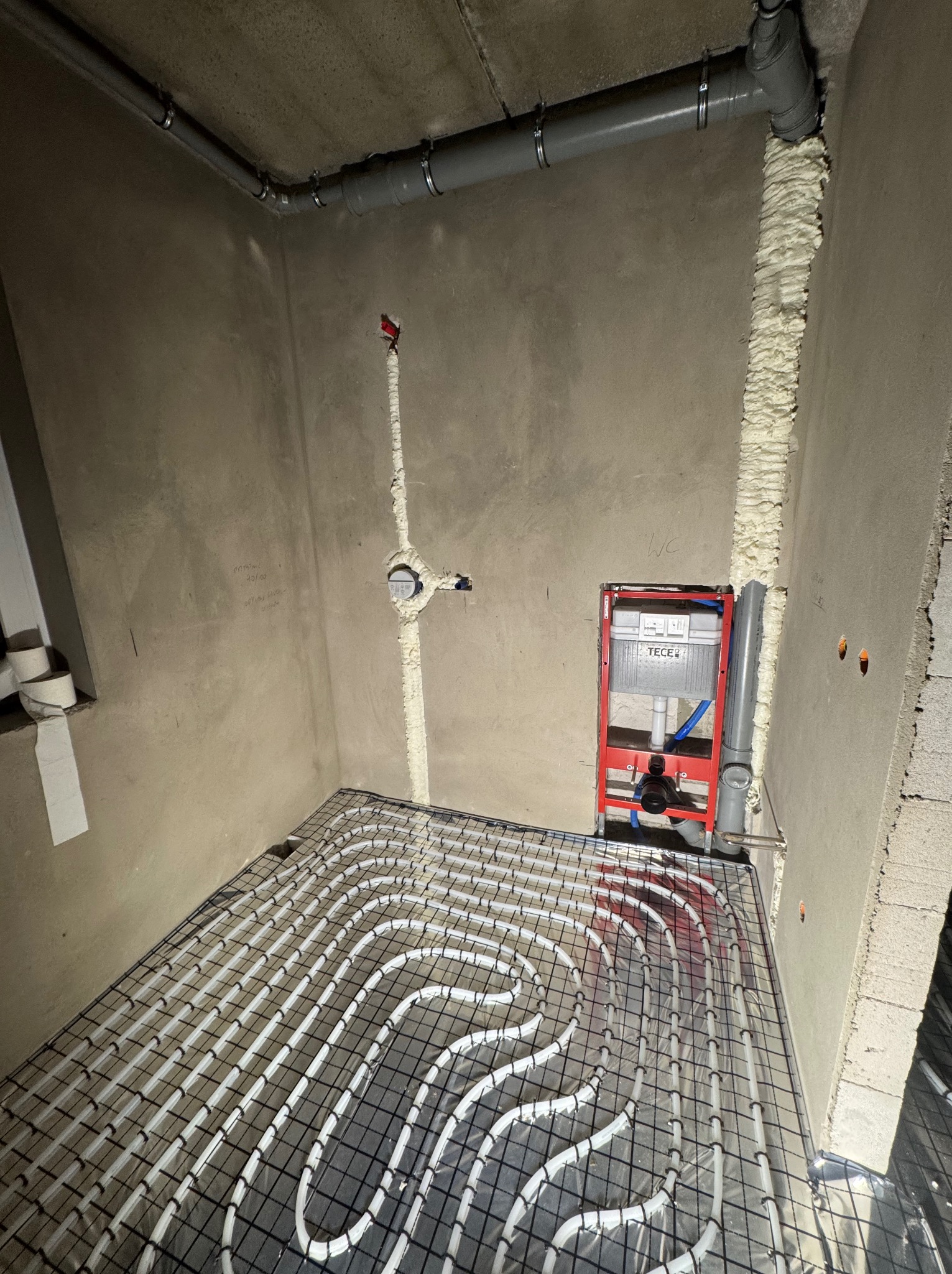 Instalacja ogrzewania podłogowego z rurami w kształcie meandrów na siatce, widoczny stelaż WC TECE, rury kanalizacyjne i ściany w surowym stanie.