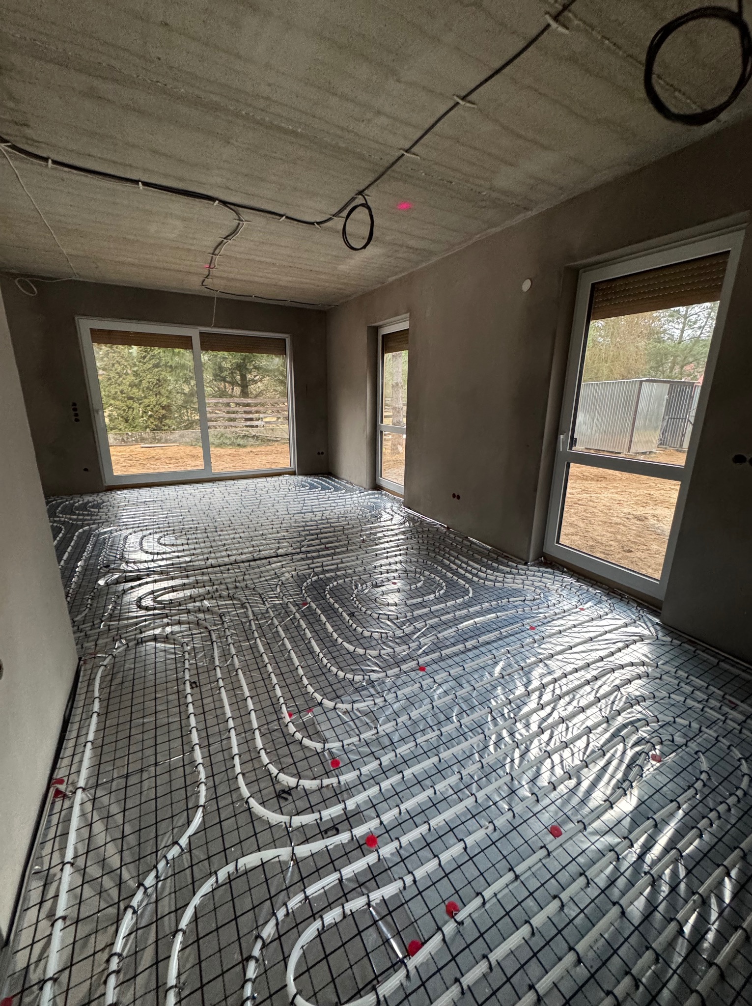 Białe rury ogrzewania podłogowego ułożone na czarnej siatce w surowym wnętrzu z oknami. Widoczne elementy instalacji na suficie. Etap przed wylewką.