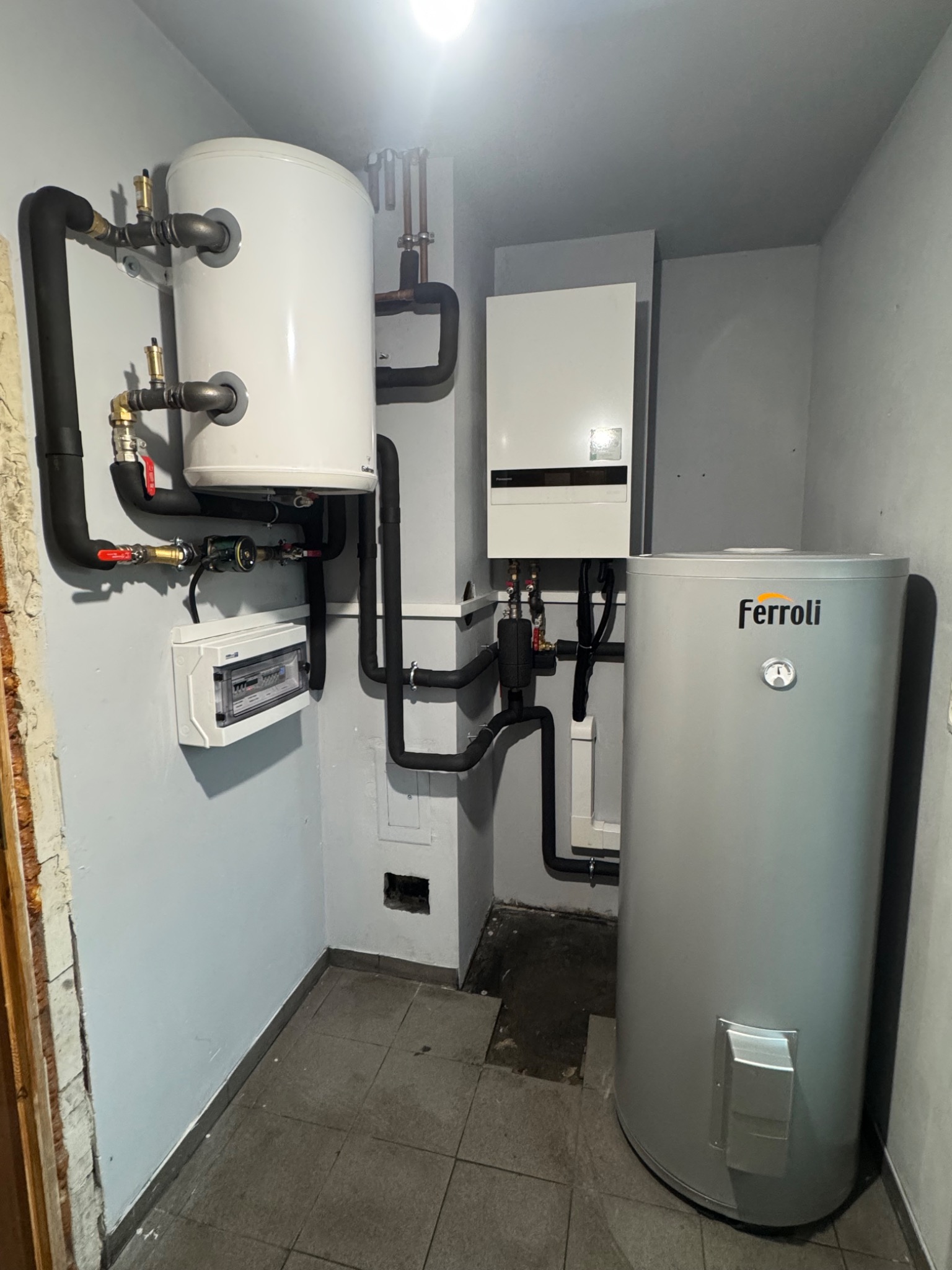 Zestaw urządzeń grzewczych Ferroli w kotłowni: zasobnik ciepłej wody, piec gazowy kondensacyjny i rozdzielnica elektryczna z czarnymi rurami instalacji CO.