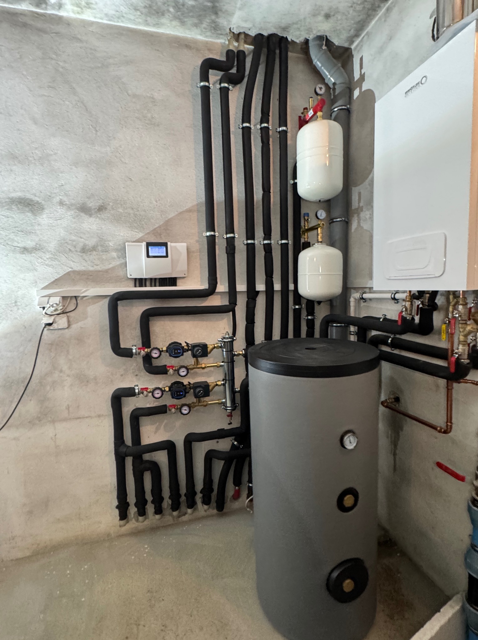 Złożona instalacja grzewcza z czarnymi rurami, białymi zbiornikami wyrównawczymi i szarym zasobnikiem ciepłej wody na tle surowej, betonowej ściany.