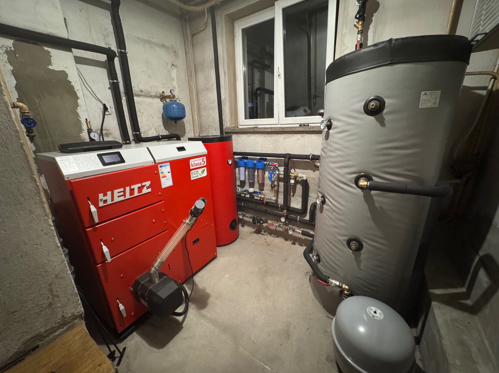 Czerwony piec CO marki HEITZ z szarym zasobnikiem wody w kotłowni. Widoczne rury, filtry i okno. Oświetlenie sufitowe. Betonowa podłoga i ściany.