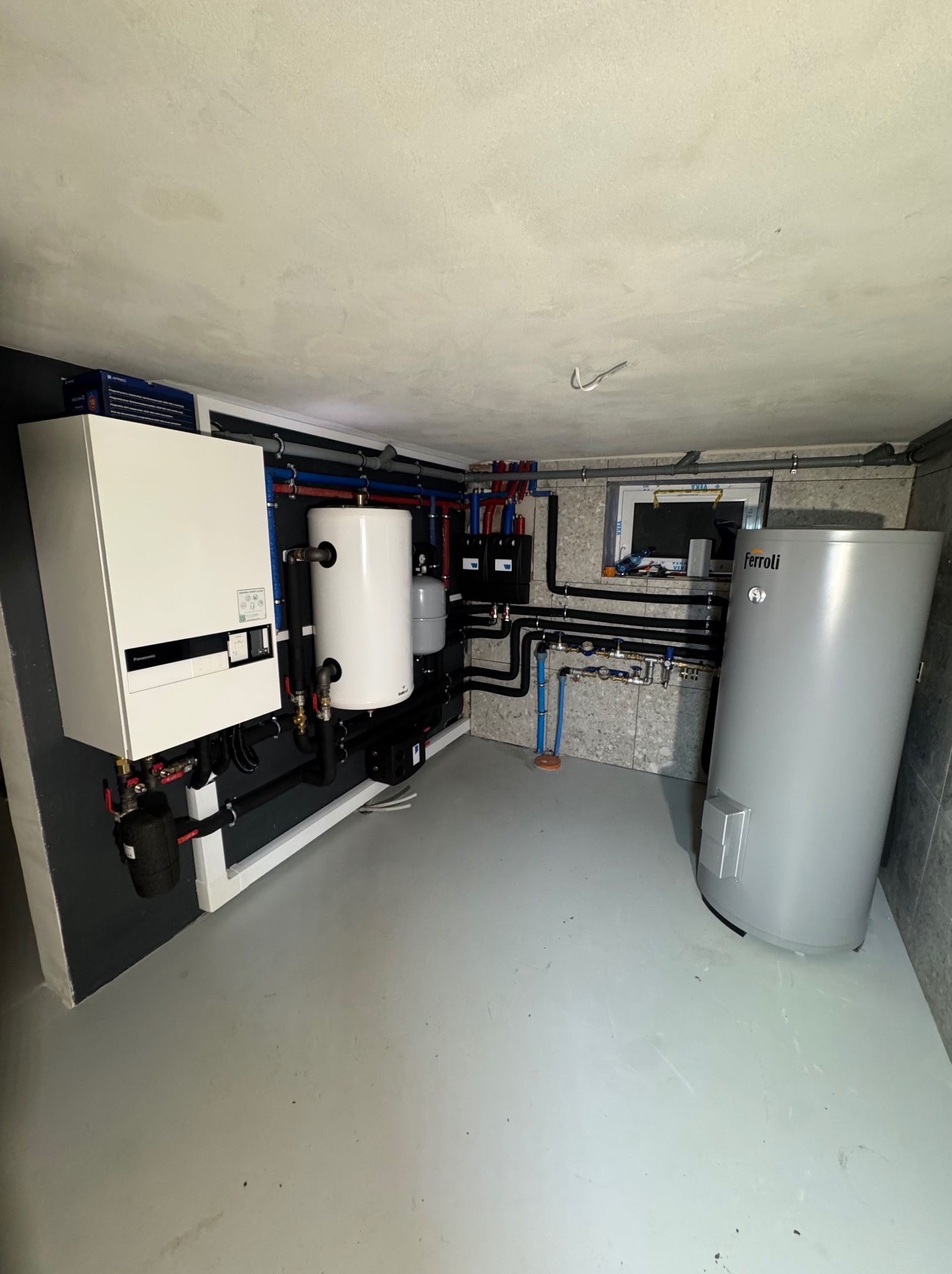 Zestaw urządzeń grzewczych: piec, zbiornik buforowy i zasobnik ciepłej wody użytkowej Ferroli. Instalacja w kotłowni z szarymi ścianami i podłogą.