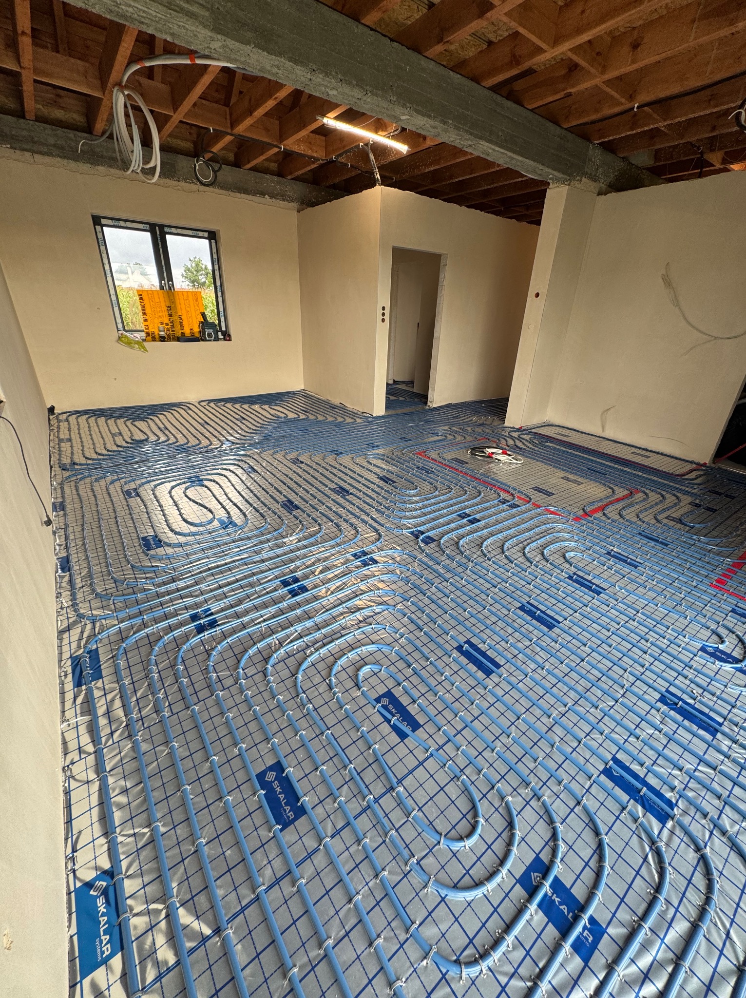 Niebieskie rury ogrzewania podłogowego ułożone na srebrnej folii w surowym wnętrzu z drewnianym stropem. Widoczne okno i instalacje elektryczne.