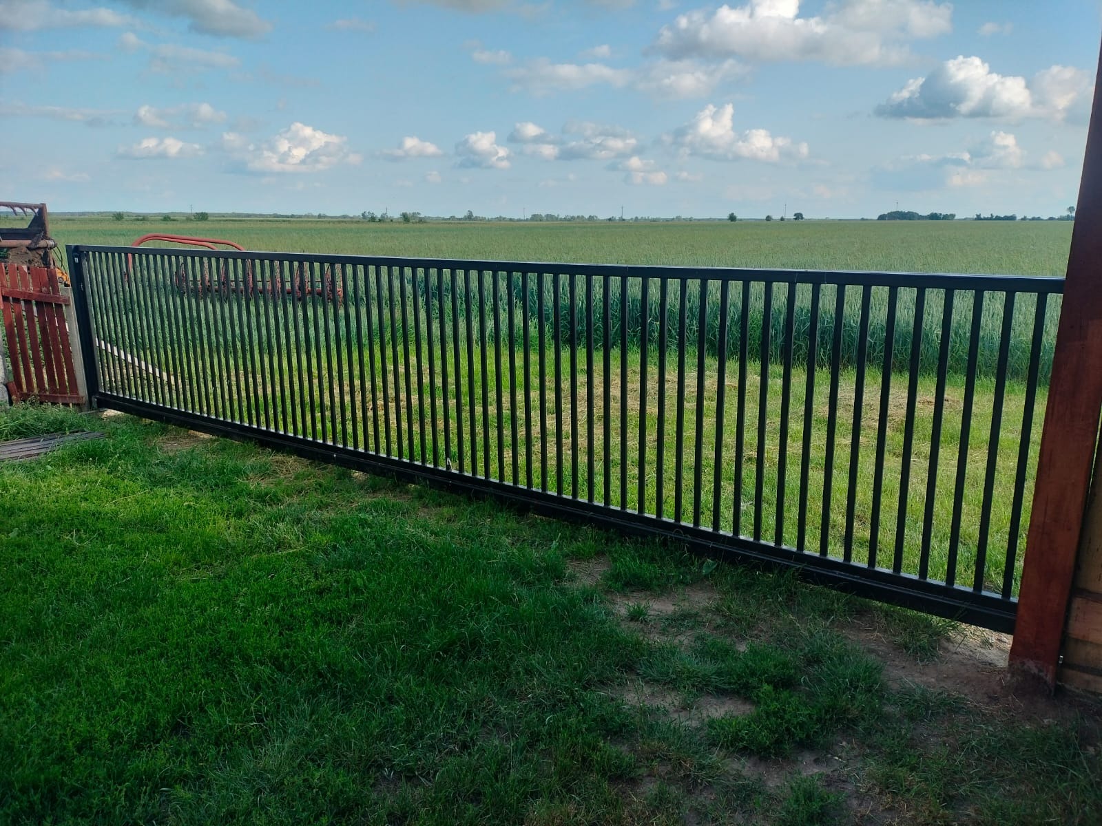 Czarna, metalowa brama przesuwna z pionowymi szczebelkami, na tle zielonego pola i błękitnego nieba z chmurami. Widoczna trawa i fragment drewnianego ogrodzenia.