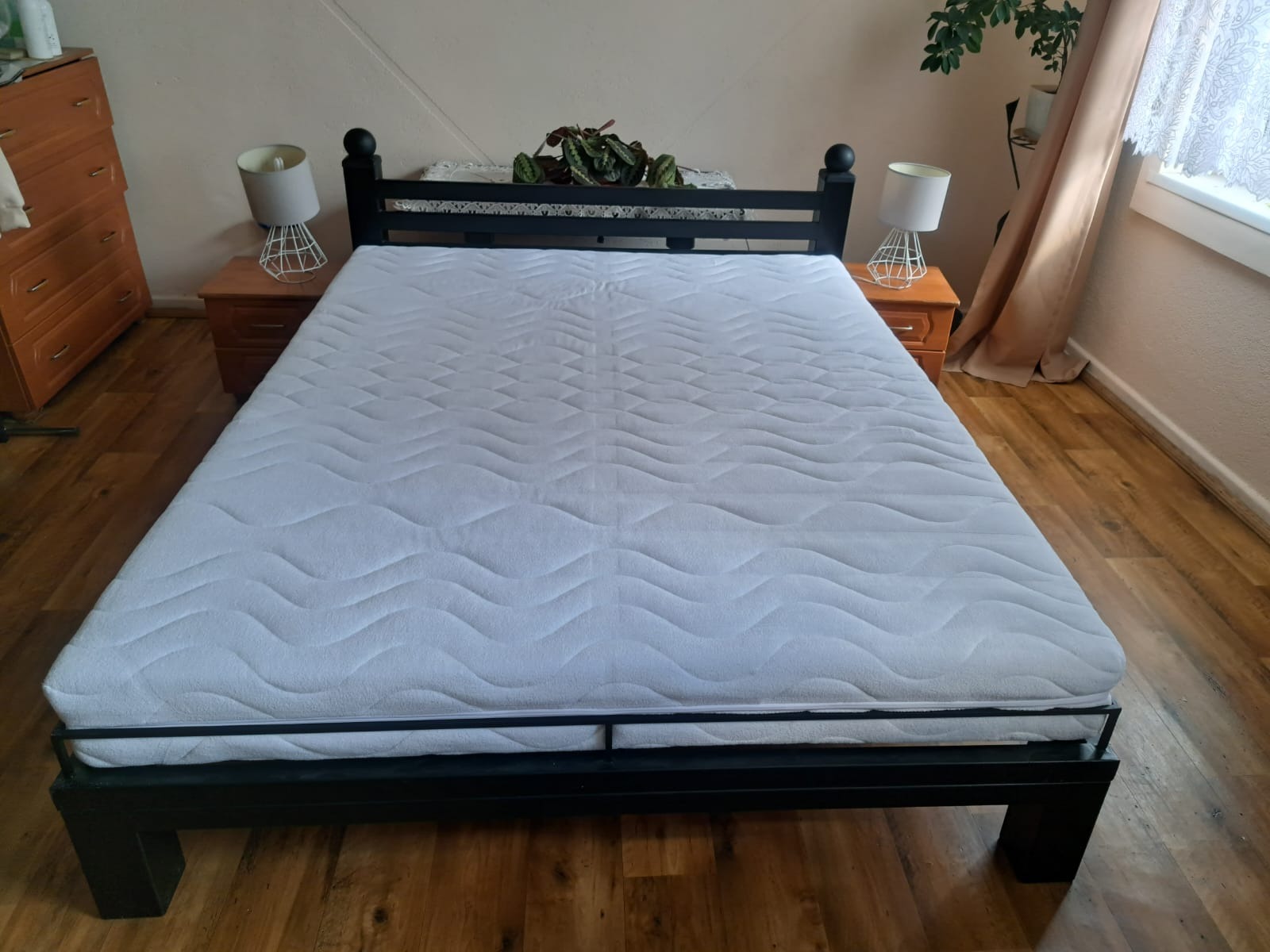 Czarne, metalowe łóżko z białym materacem w sypialni z drewnianą podłogą. Widoczne szafki nocne z lampkami, komoda i roślina doniczkowa.