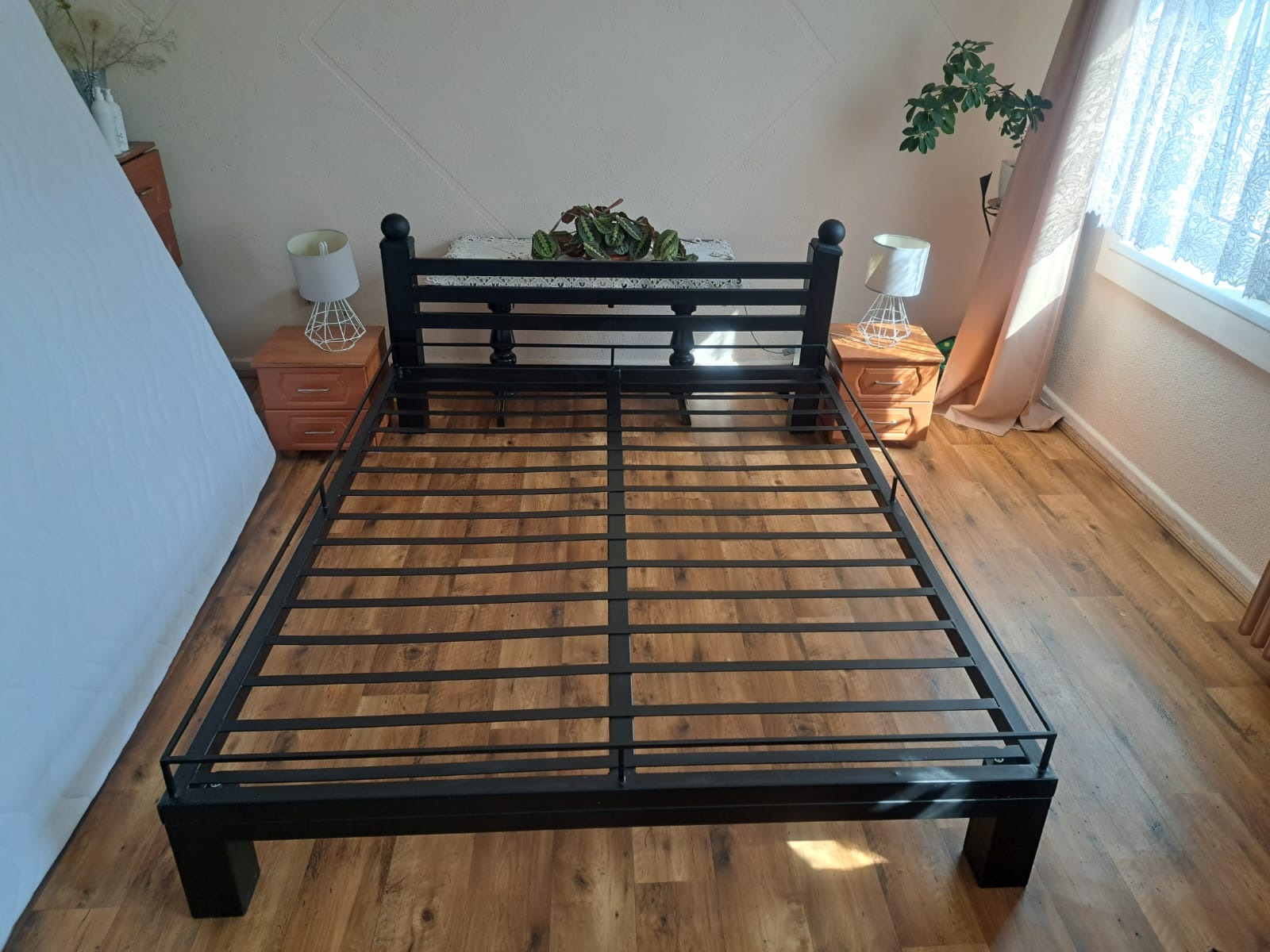 Metalowa rama łóżka z czarnymi, okrągłymi zakończeniami, ustawiona na drewnianej podłodze, z szafkami nocnymi i lampkami po bokach, w jasnym wnętrzu.