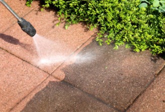 Częściowo umyta, brudna kostka brukowa z bliska, zraszacz pod ciśnieniem, zielona roślina w tle. Widoczna różnica w kolorze kostki przed i po myciu.