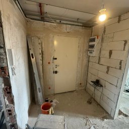 Volti Instalacje Elektryczne Michał Janczy - Remont korytarza: widok na drzwi wejściowe, ściany w trakcie wykończenia, widoczna instalacja elektryczna, poziomica i narzędzia. Żarówka oświetla wnętrze.
