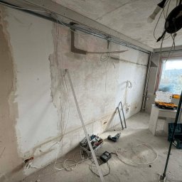 Volti Instalacje Elektryczne Michał Janczy - Wnętrze w trakcie remontu: widoczne przewody elektryczne pod sufitem, ściany przygotowane do tynkowania, narzędzia i skrzynki narzędziowe na podłodze. Etap prac elektrycznych.