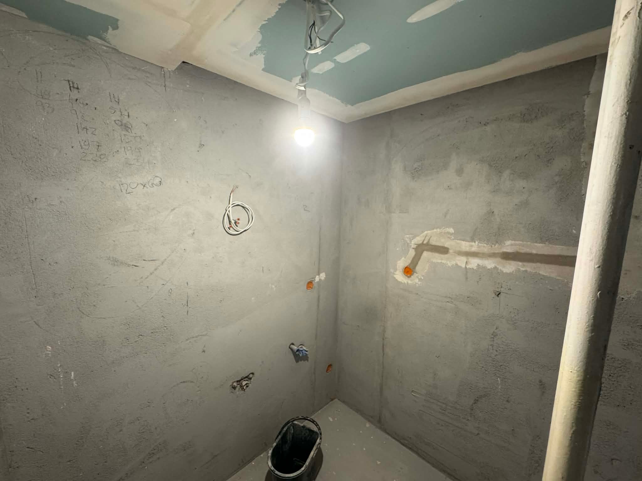 Surowy stan łazienki w trakcie remontu: widoczne instalacje elektryczne i hydrauliczne w ścianach, sufit z płyt gipsowych, wisząca żarówka i wiadro na podłodze.