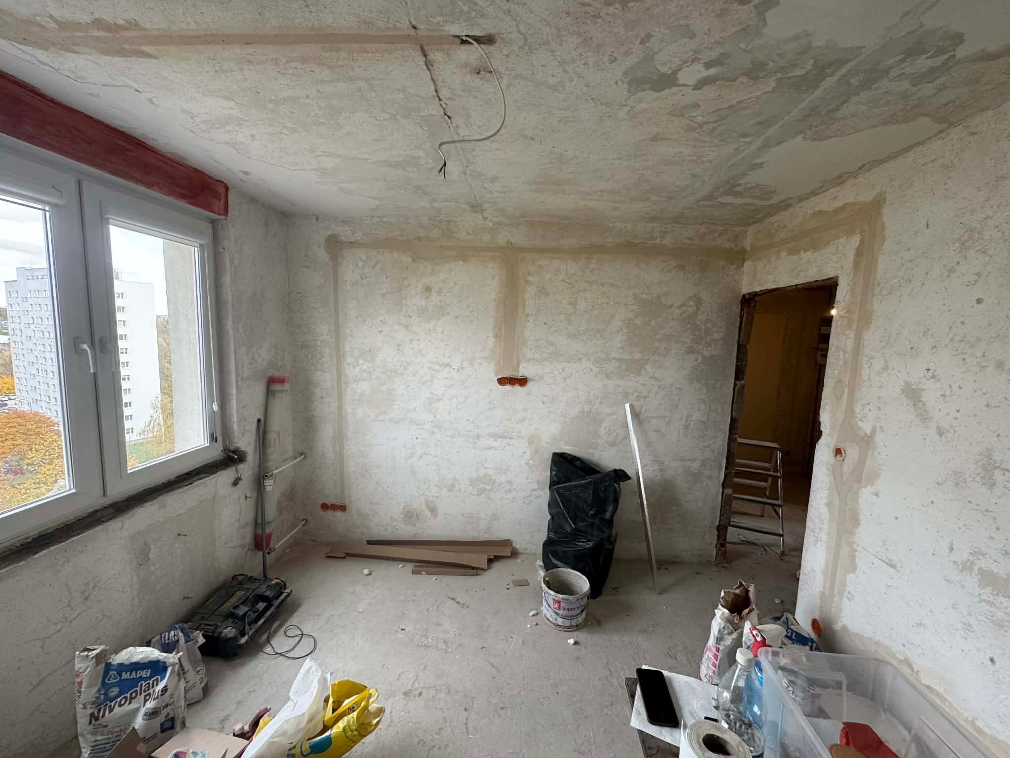 Wnętrze w trakcie remontu: surowe ściany z widocznymi przewodami elektrycznymi, okno z widokiem na drzewa, narzędzia i materiały budowlane. Etap prac wykończeniowych.