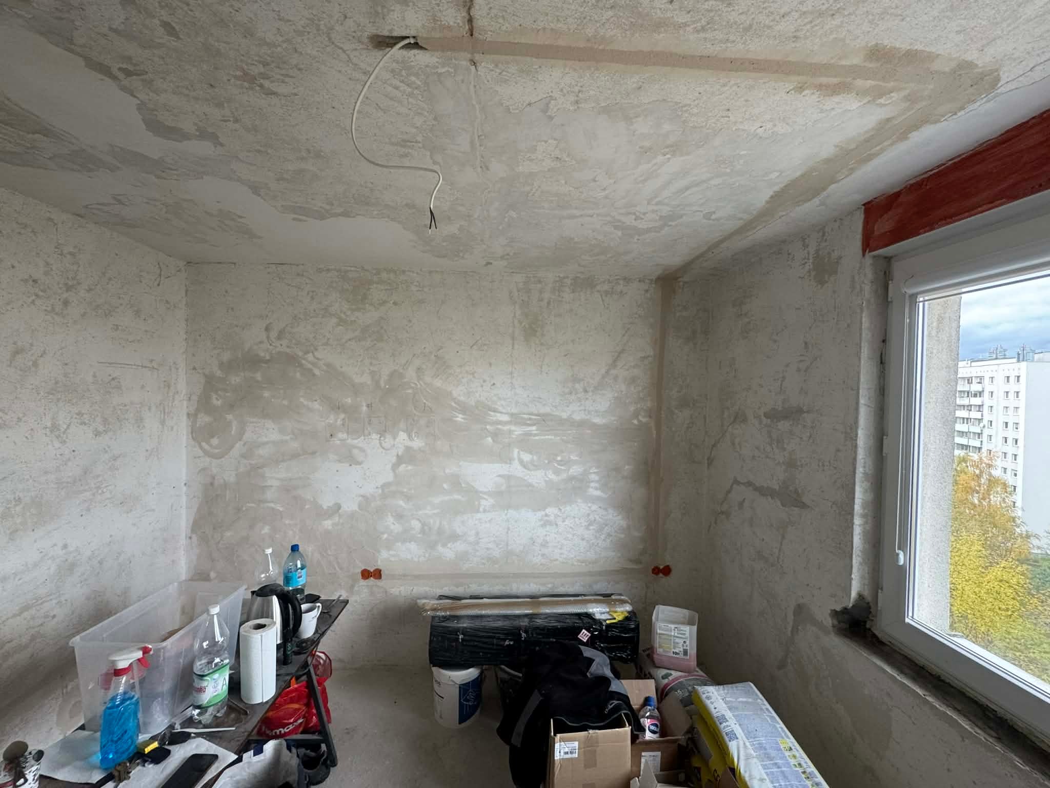 Surowe wnętrze w trakcie remontu: widoczne ślady po instalacji elektrycznej na suficie, przygotowane ściany do malowania i okno z widokiem na zewnątrz.