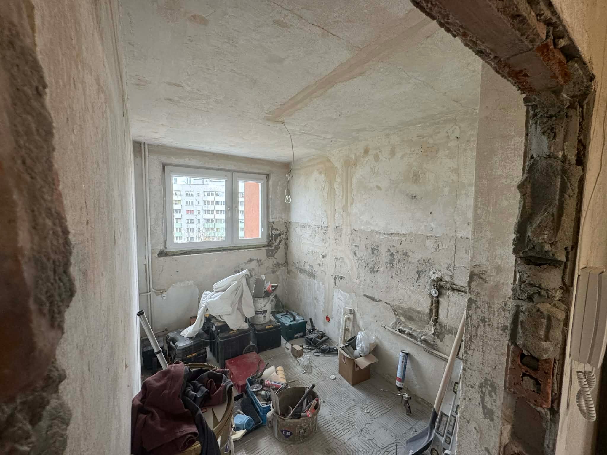 Pokój w trakcie remontu: surowe ściany, odsłonięta instalacja, narzędzia i materiały budowlane. Widoczne ślady po kuciu i przygotowaniu do dalszych prac.