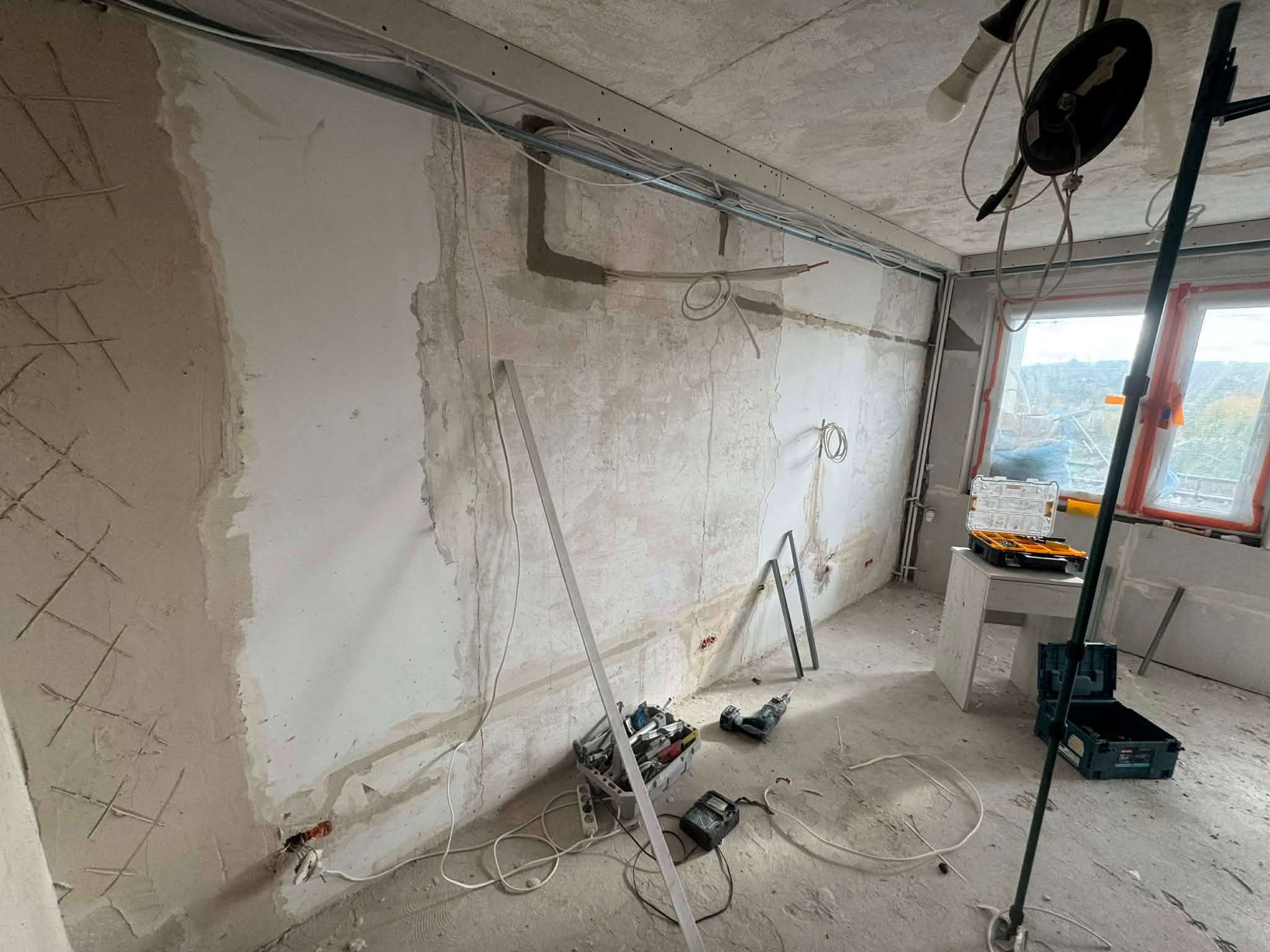 Wnętrze w trakcie remontu: widoczne przewody elektryczne pod sufitem, ściany przygotowane do tynkowania, narzędzia i skrzynki narzędziowe na podłodze. Etap prac elektrycznych.