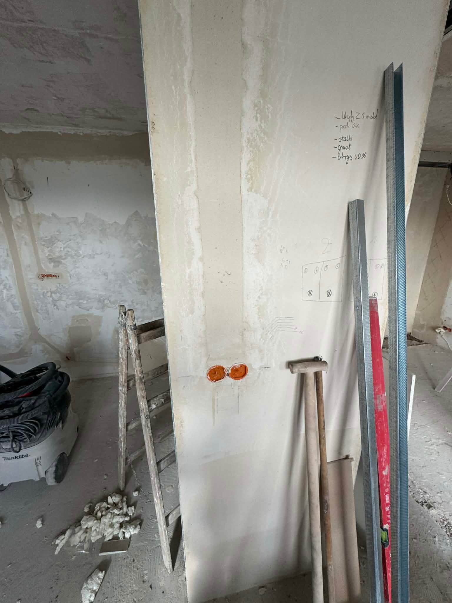 Wnętrze w trakcie remontu: widoczne puszki elektryczne w ścianie z karton-gipsu, profile metalowe, drabina i odkurzacz. Na ścianie notatki i schemat instalacji.