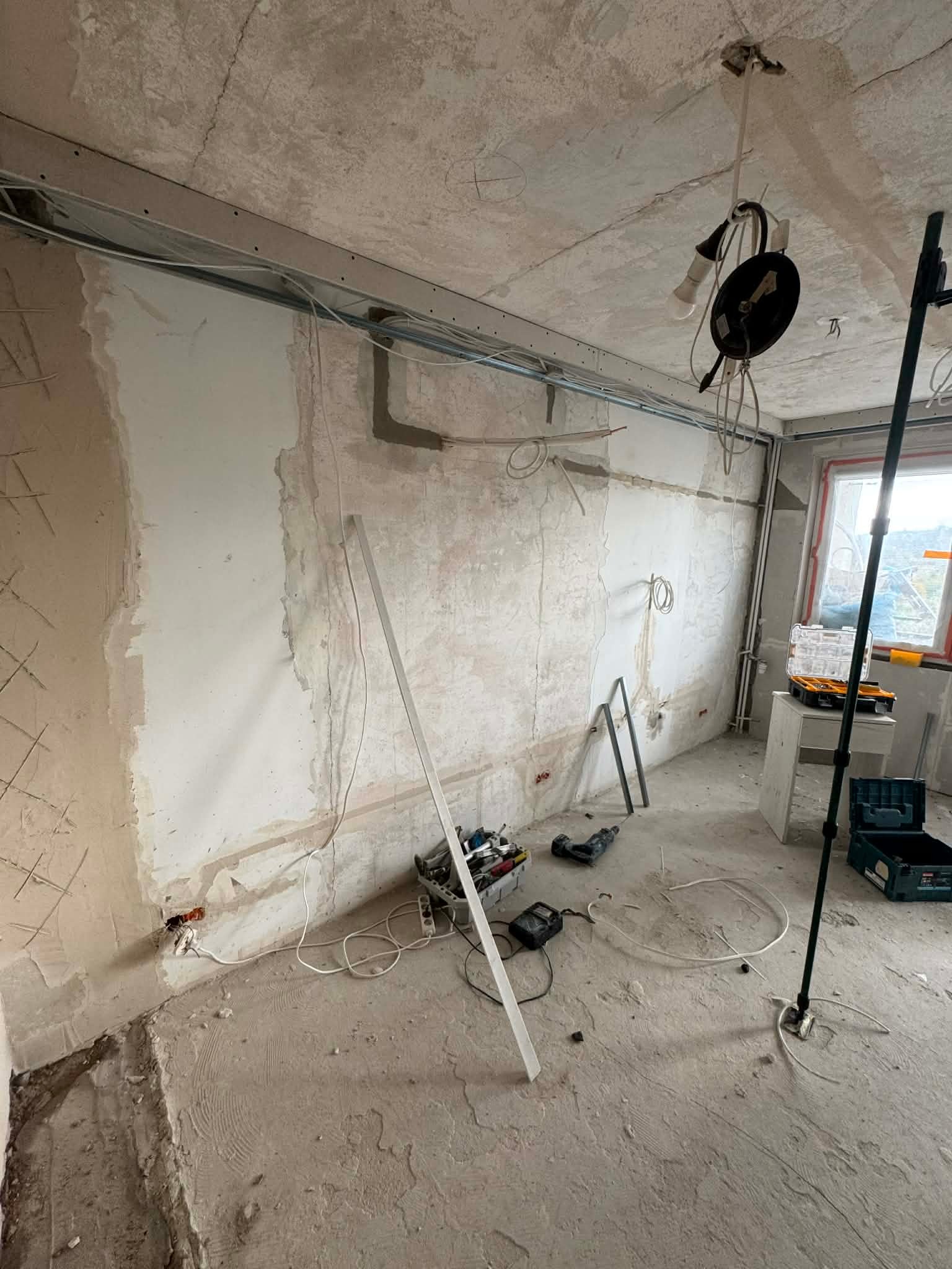 Remontowany pokój z widoczną instalacją elektryczną pod sufitem, narzędziami i przewodami na podłodze. Ściany w trakcie przygotowania do malowania.